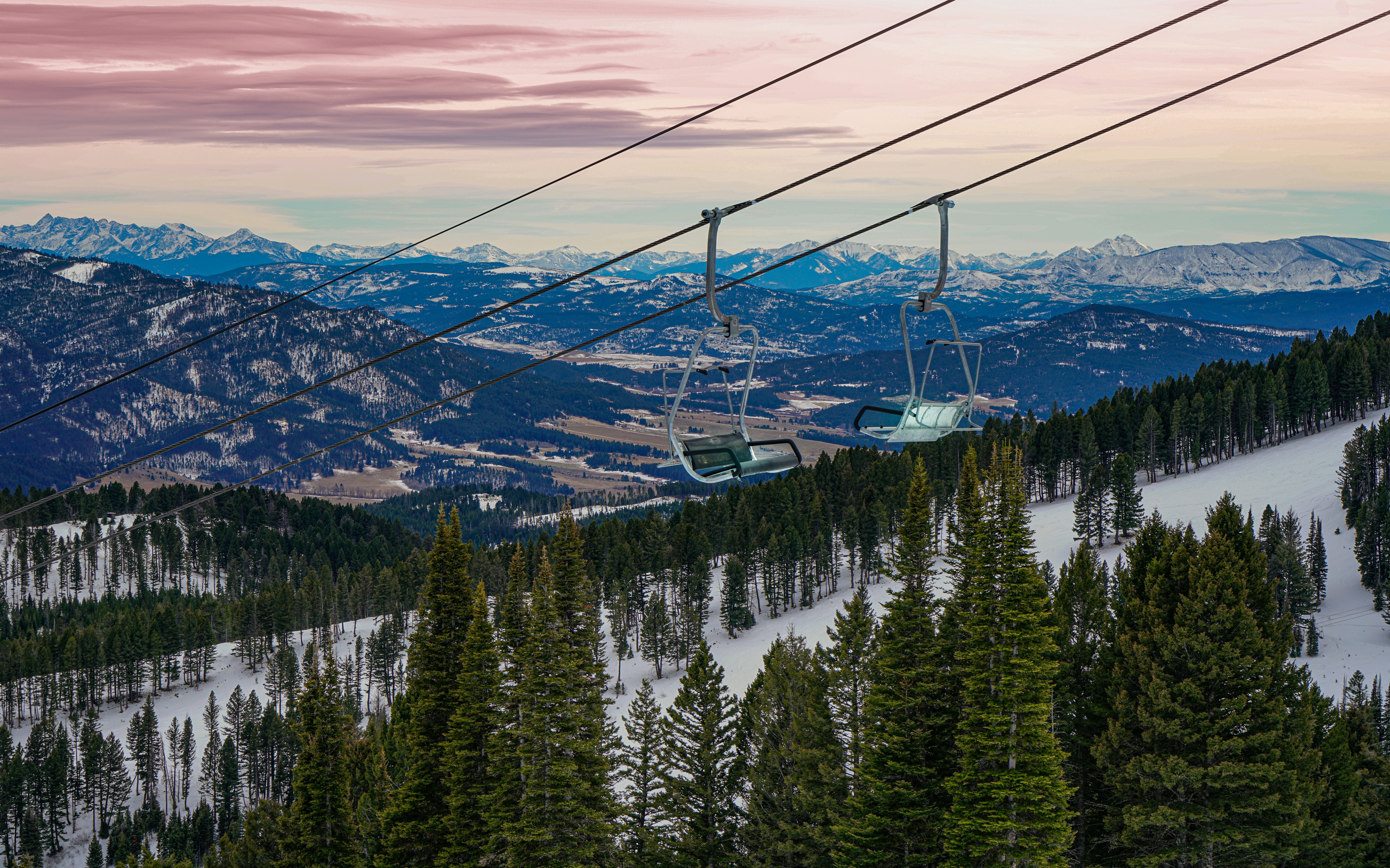 A ski lift goes up the mountain with a valley behind and pink skies
