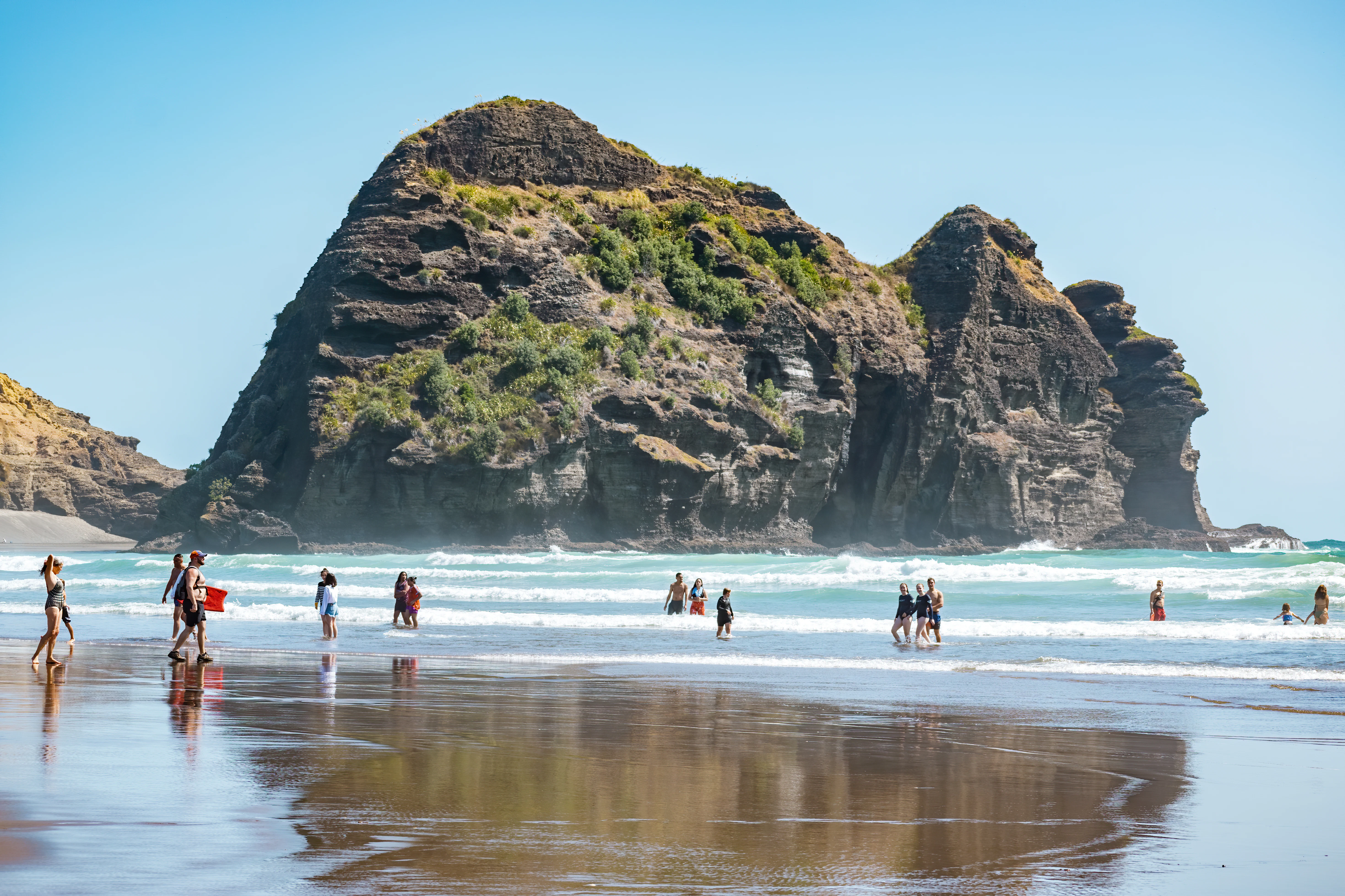 People swimming and walking at a beach in Auckland, New Zealand, with a large rock formation in the water.