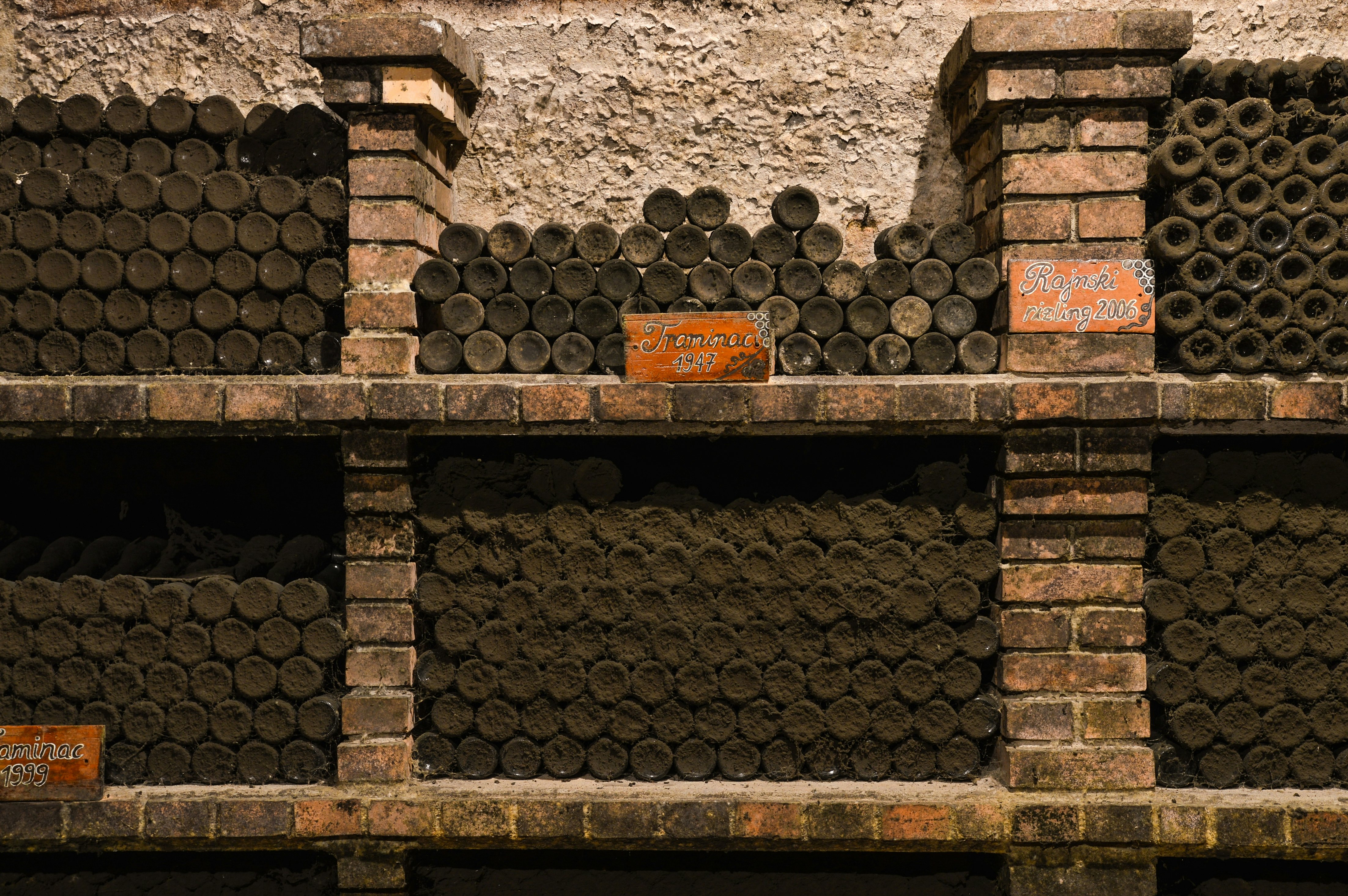 Dark, dusty wine bottles are stacked on their sides so only the bottoms are visible between brick shelves.