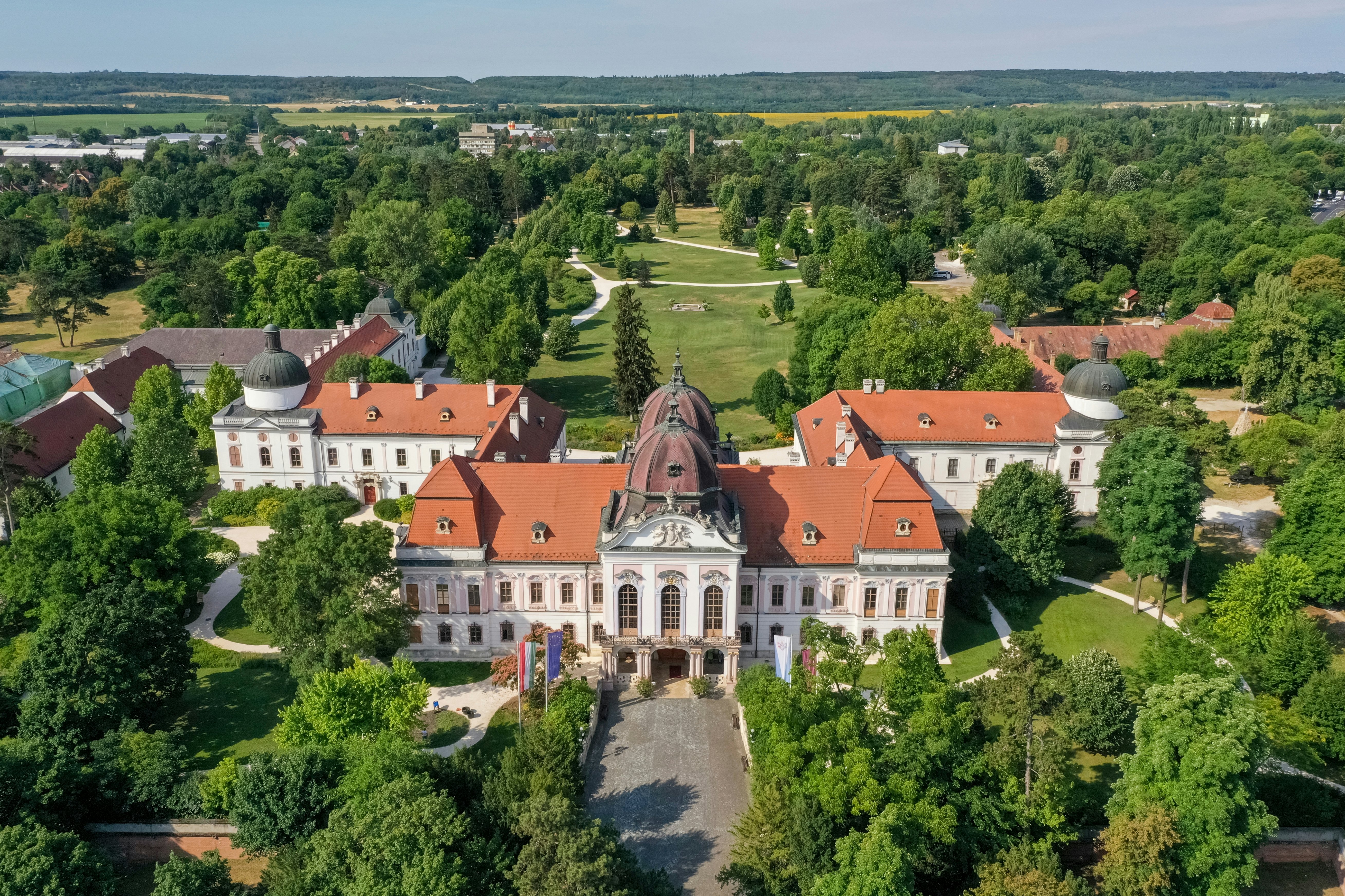 An aerial view of a white palace with a red roof set in grounds dense with green trees.