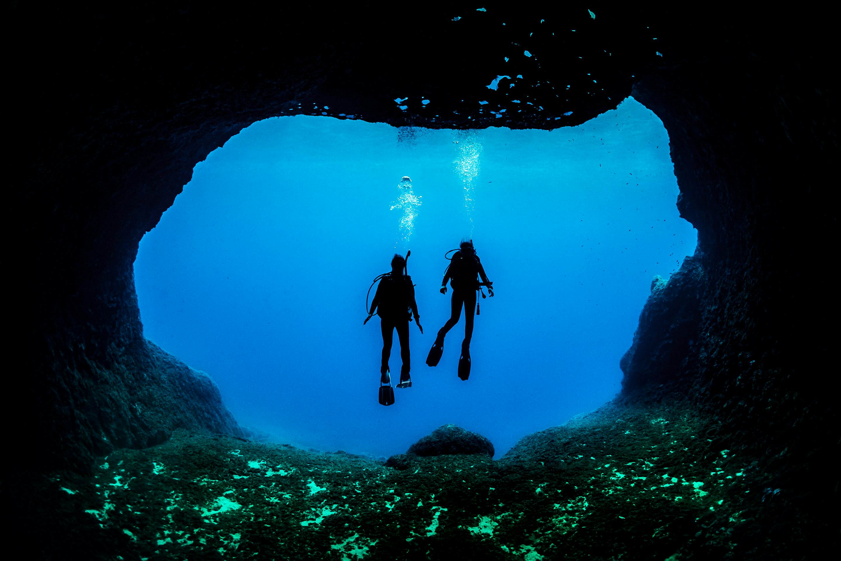 Silhouettes of two people in scuba gear underwater at the opening of a cave.