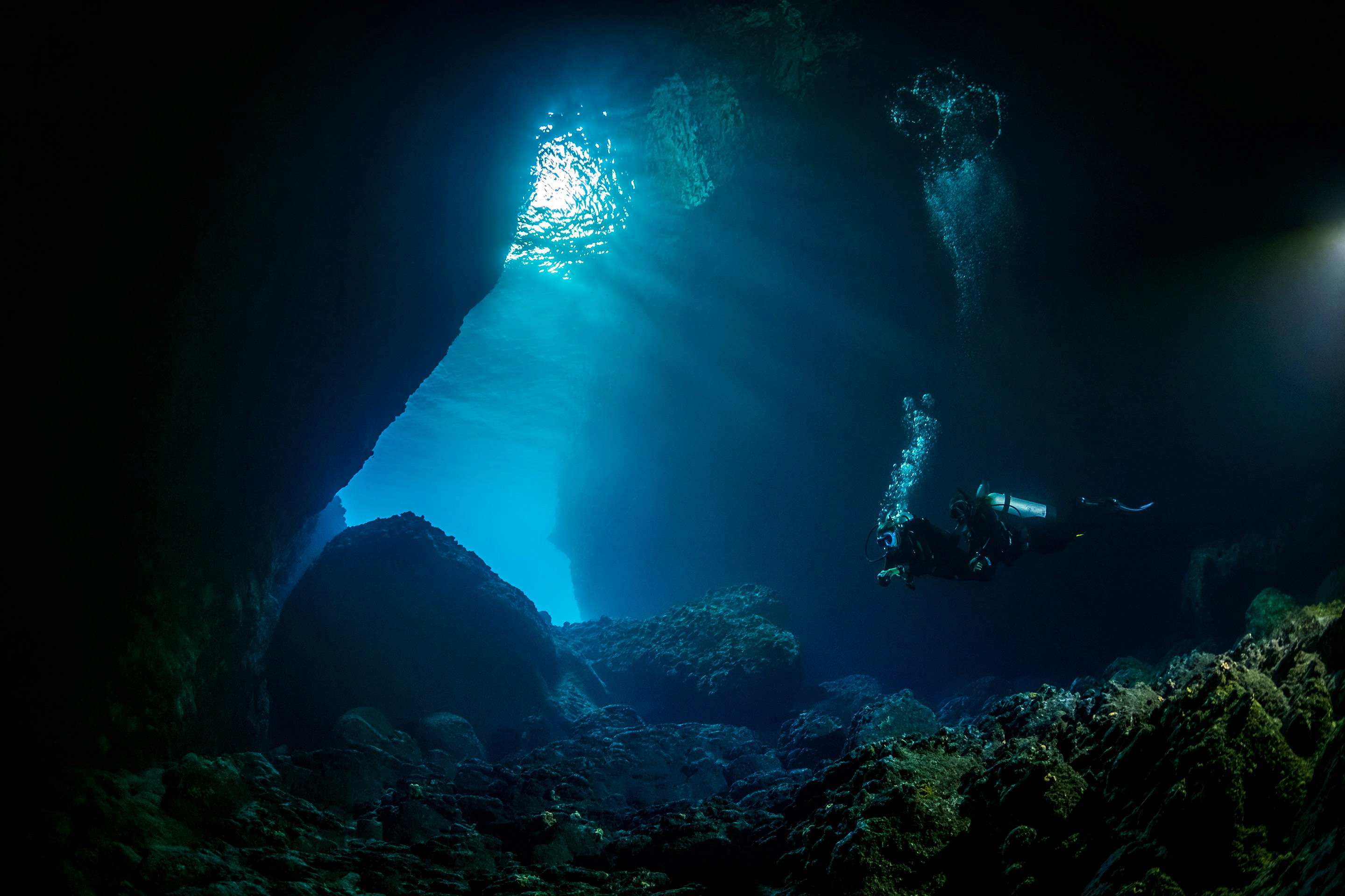 Two people scuba diving in a cave, with light shining through a hole in the cave