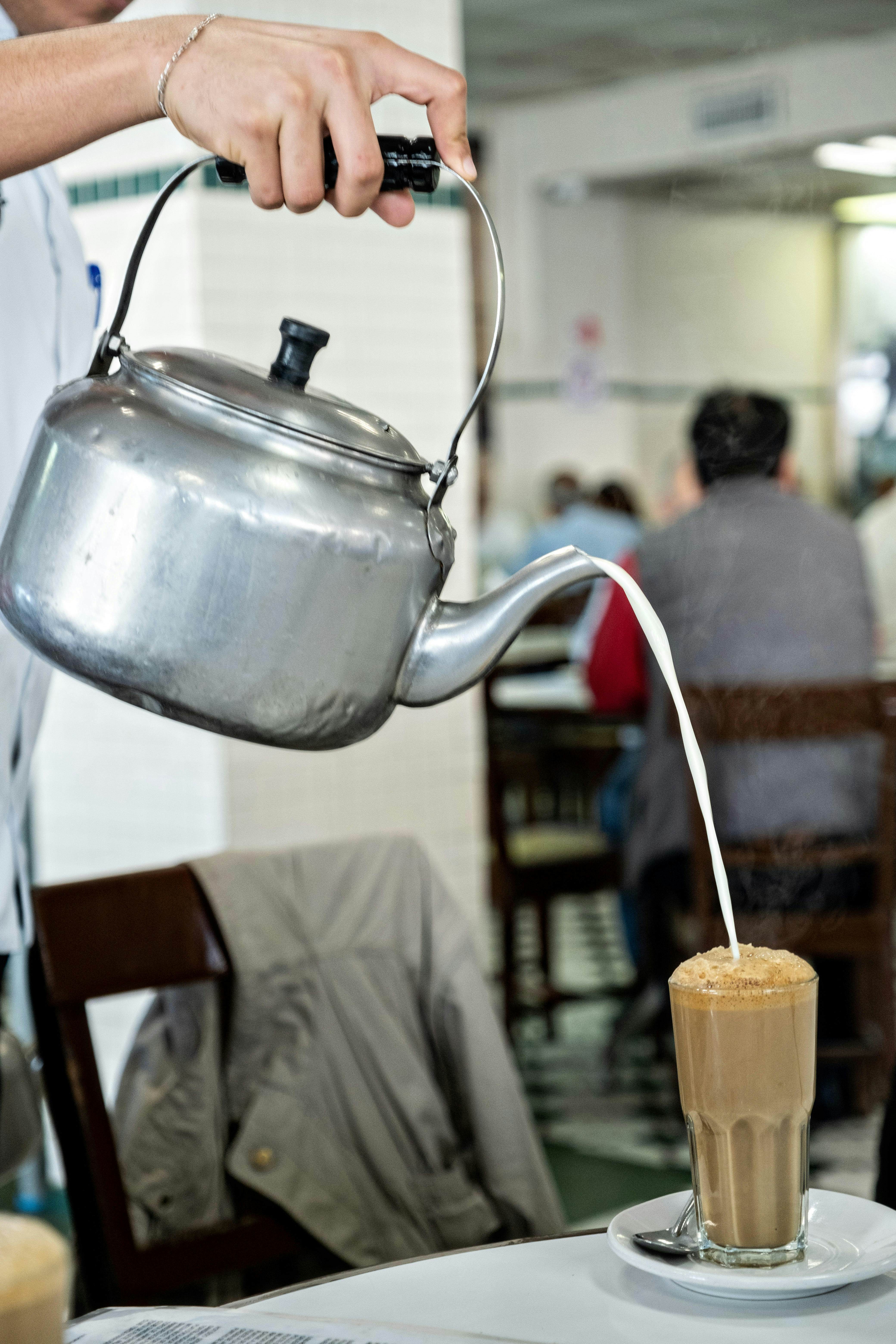 A waiter pours hot milk from a spouted silver kettle into coffee in a tall, clear glass.