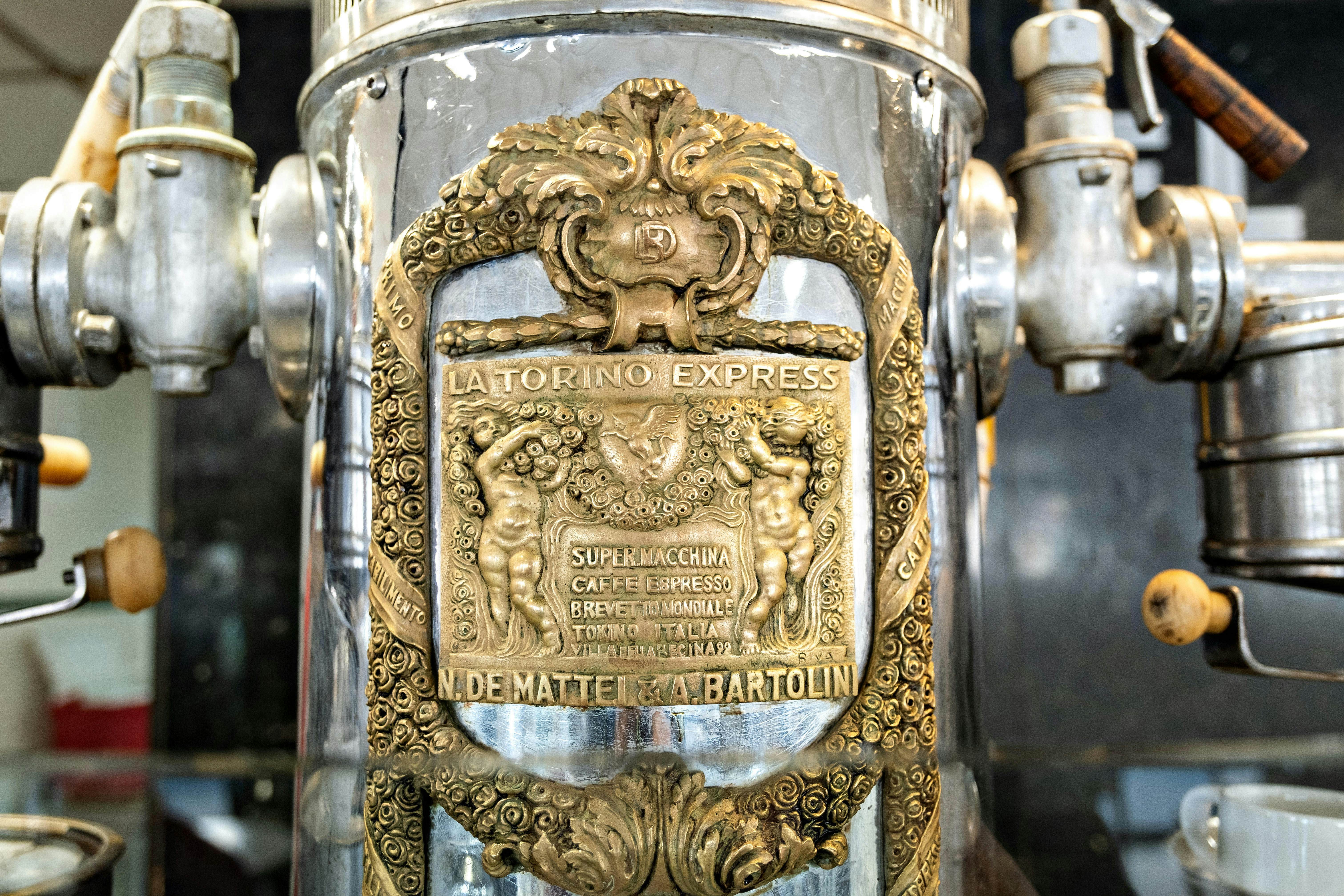 Close-up of old-style espresso machines with a bronze-colored plaque on the front of one reading "La Torino Express."