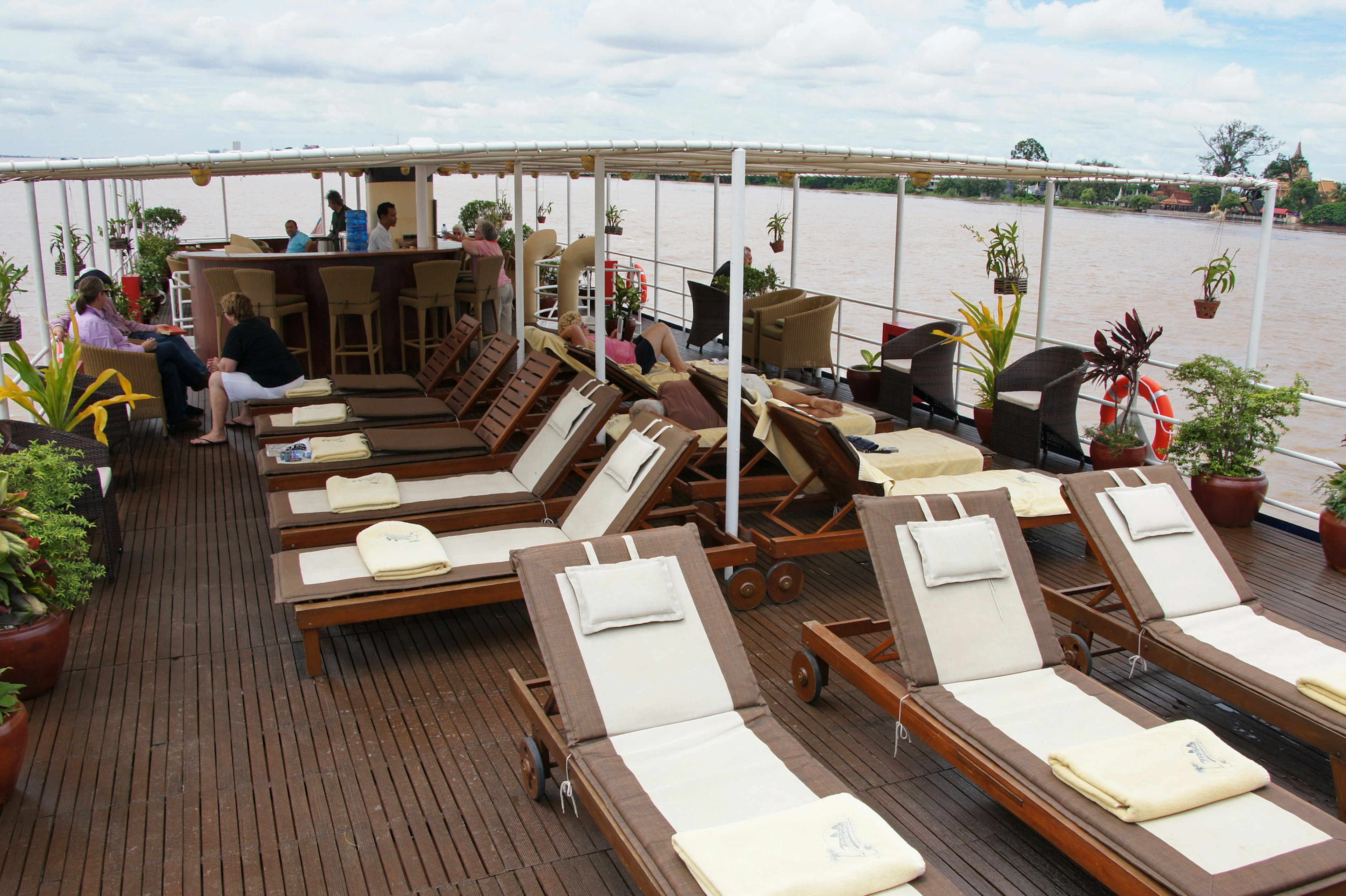 People relax in wicker chairs and on sunloungers on the covered wooden deck of a boat as it travels down a river.