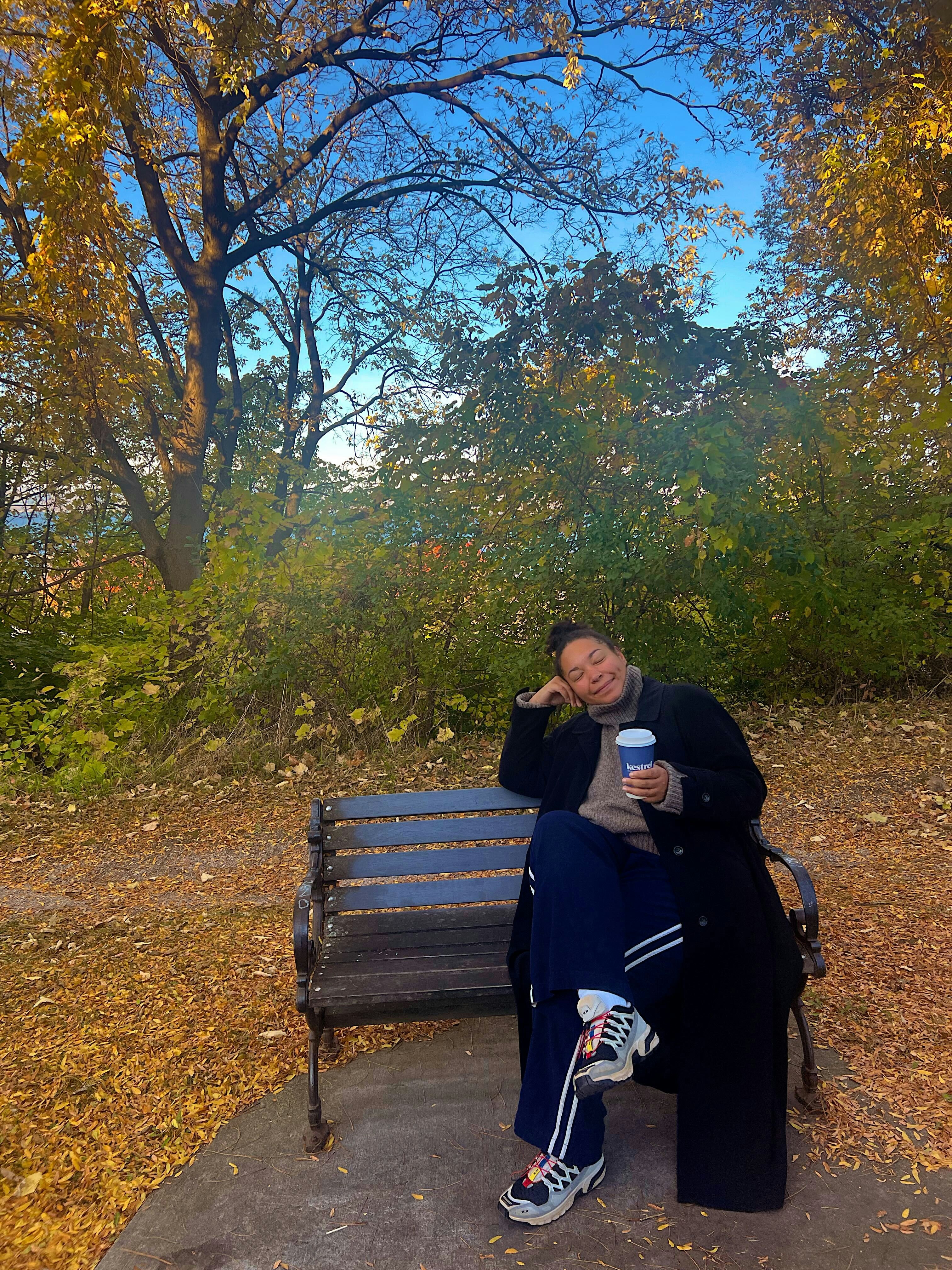 A woman wearing a sweater and long coat and holding a paper cup of coffee sits on a bench surrounded by golden leaves.