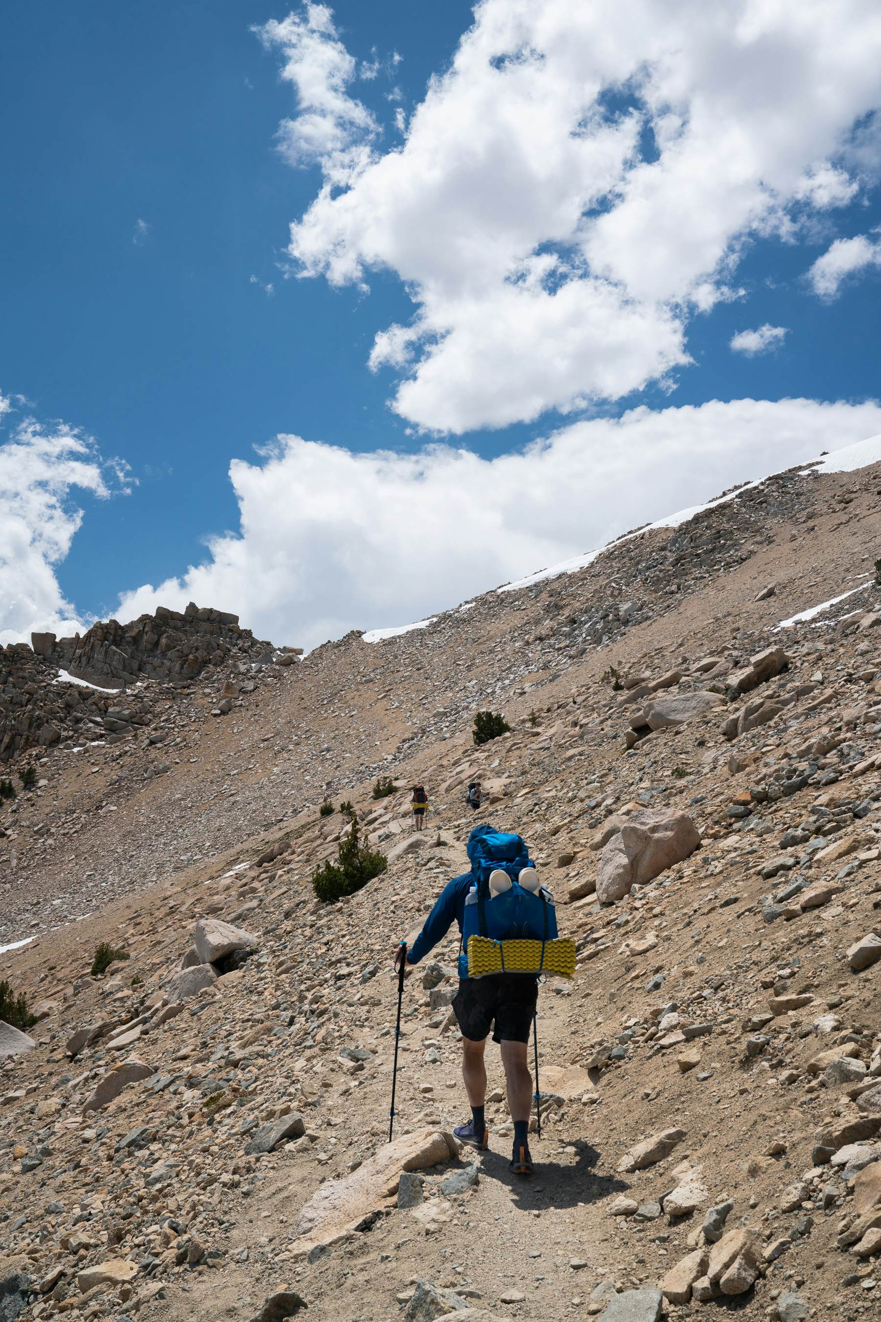 A hiker, carrying a backpack and using hiking poles, follows a rocky mountain trail.