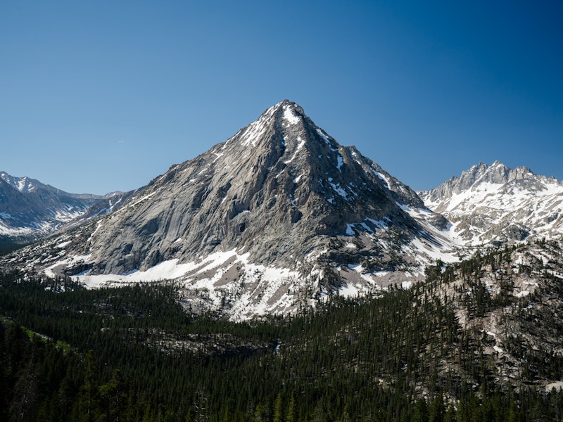 A pointed mountain peak with snow near its base where the mountain meets the tree line.