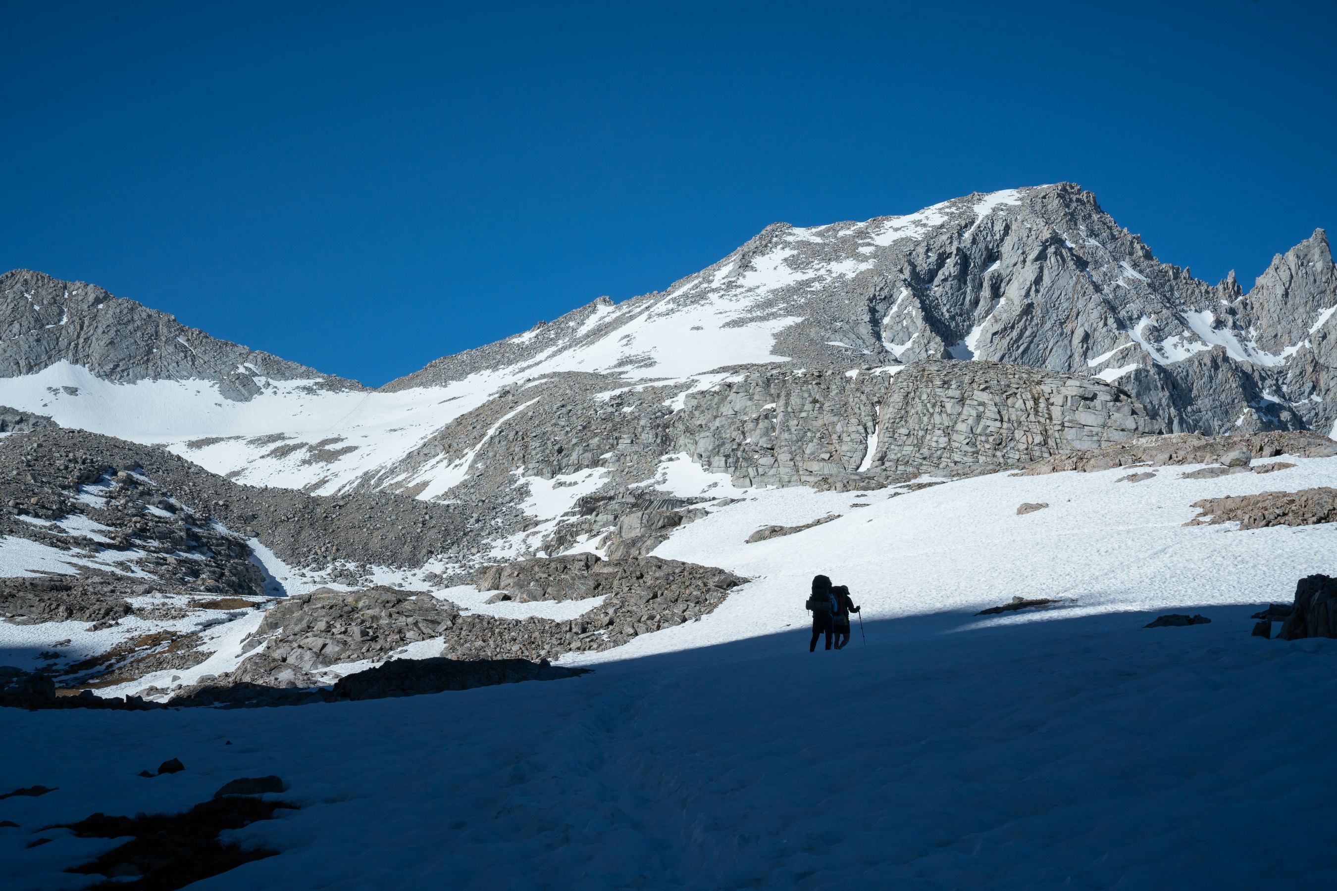 Two hikers in silhouette move from the shadow of a mountain and enter the sunlight on a snowy mountain trail.