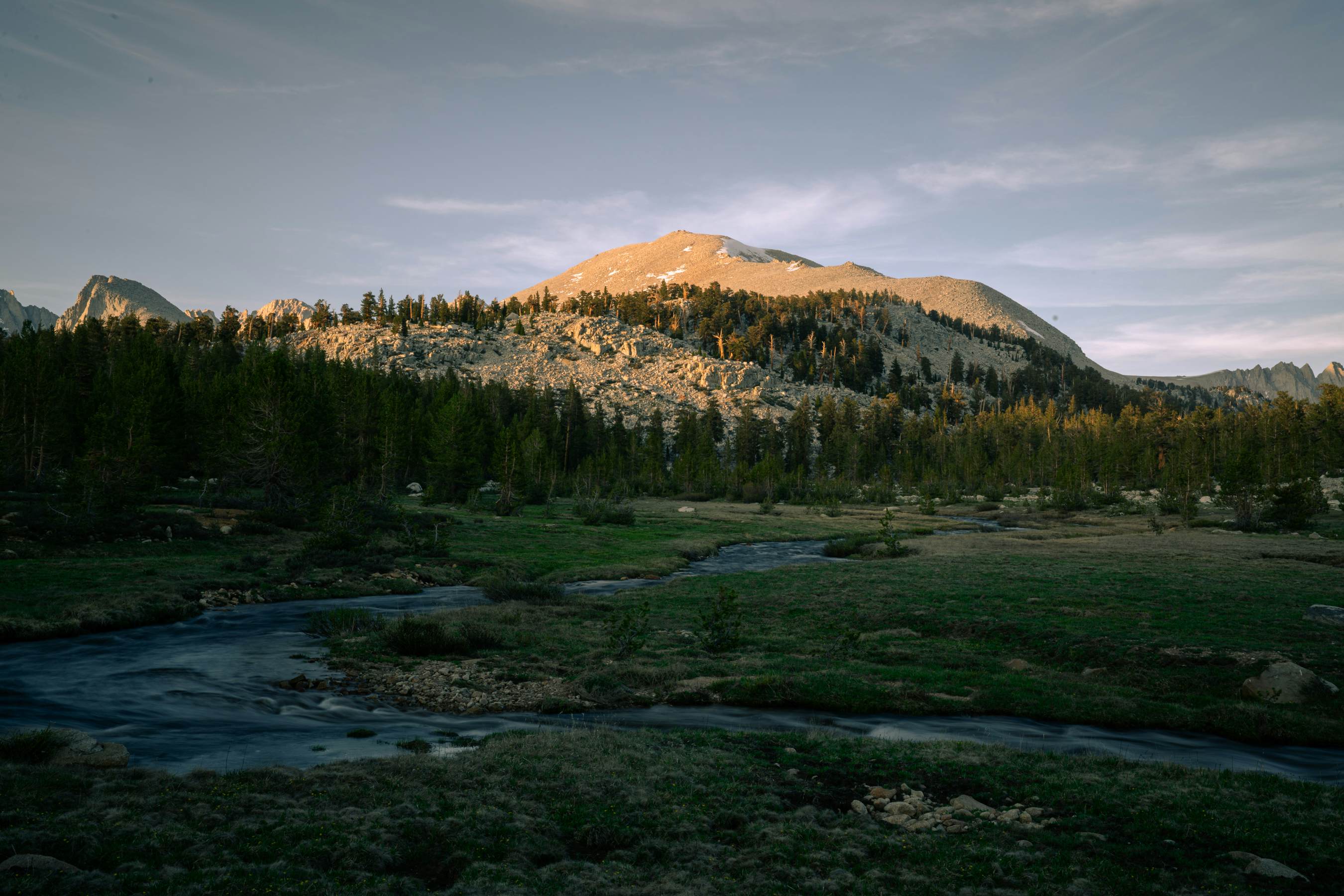 A creek weaves its way through a meadow at the foot of rolling hills covered in woodland.