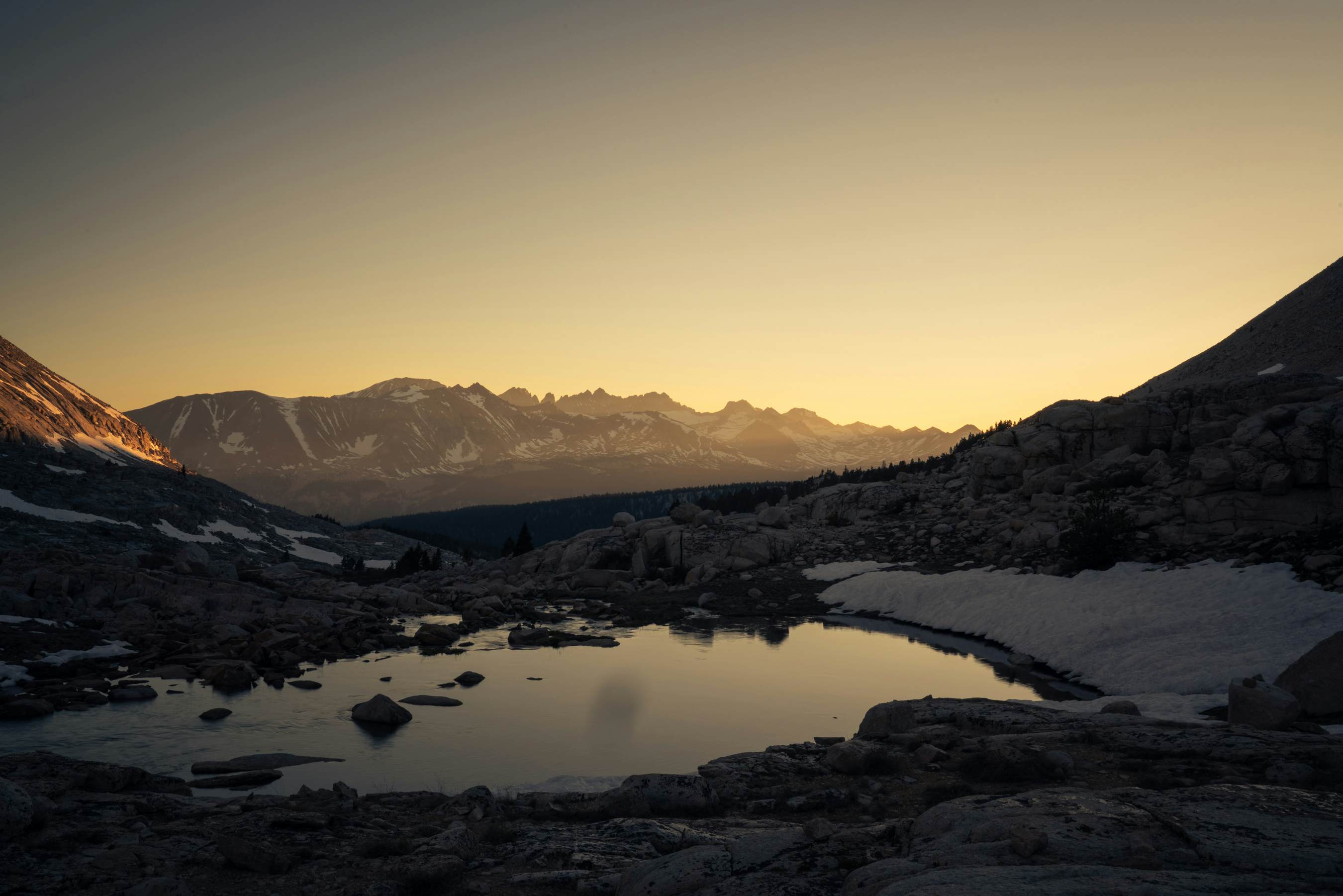 A mountain lake reflects the rising sun as the sky lights up in pale yellow.