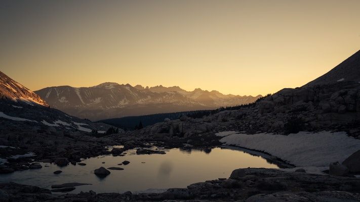 A mountain lake reflects the rising sun as the sky lights up in pale yellow.