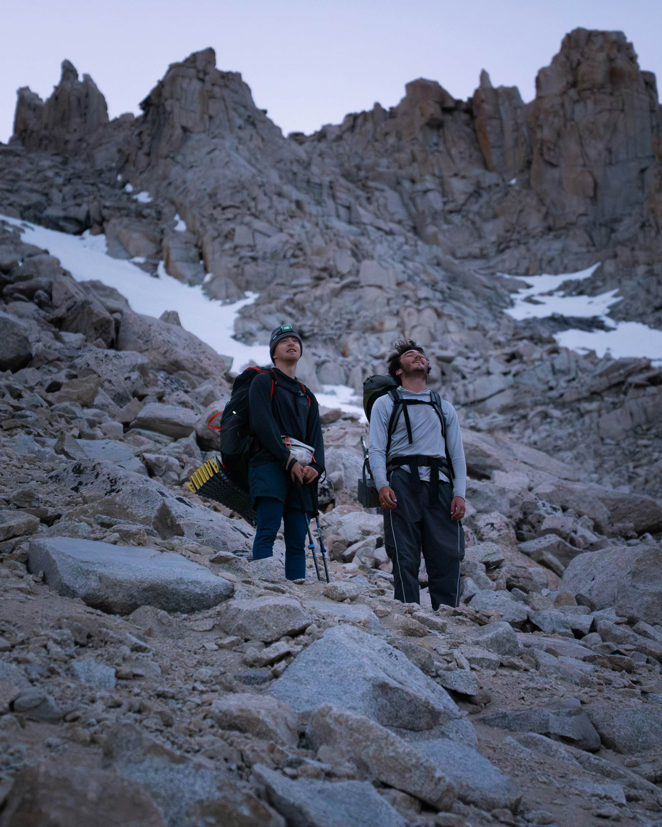 Two male hikers carrying backpacks pause on a mountain trail to watch the sunrise.