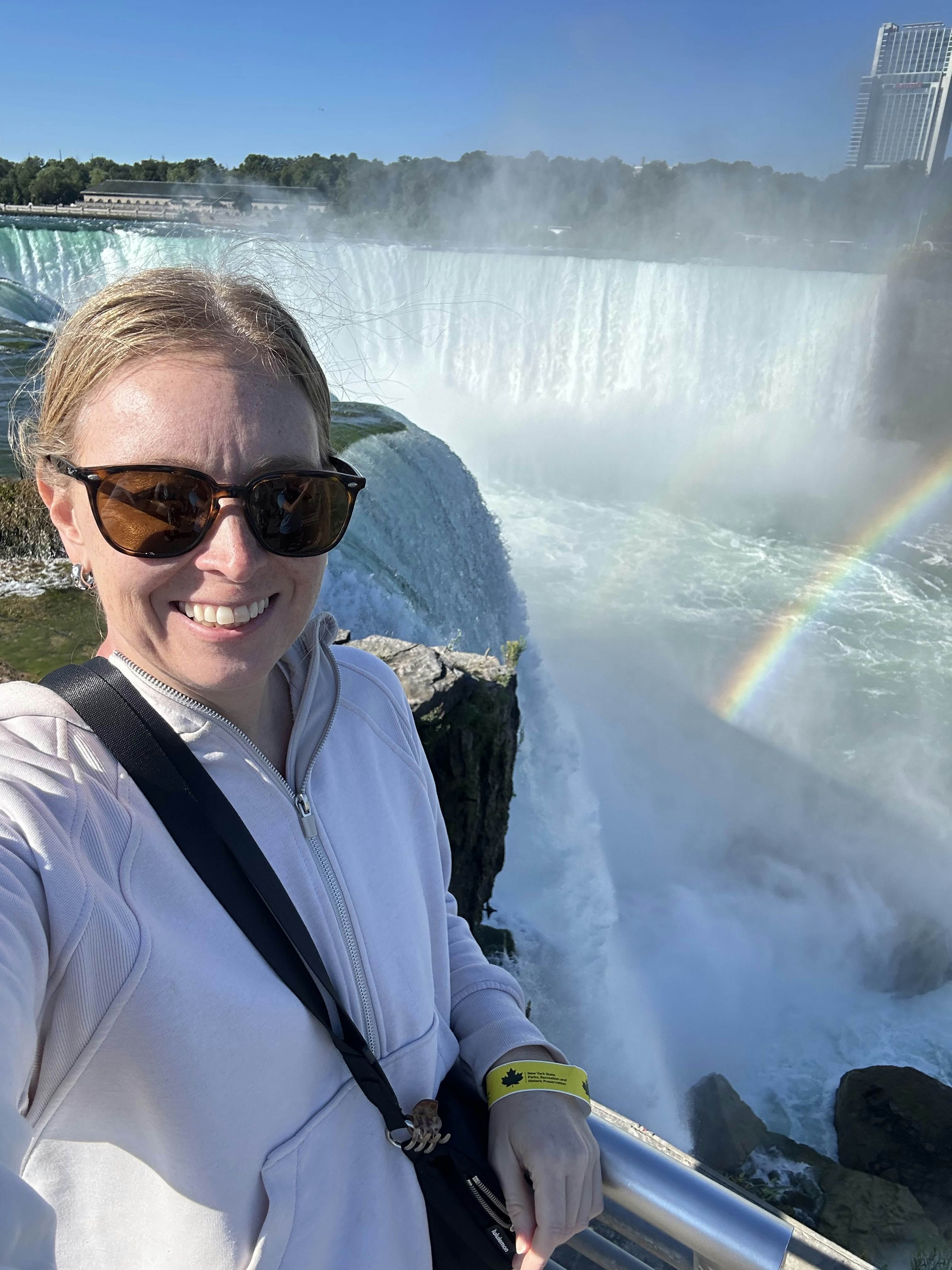 Woman in sunglasses and a tan sweater with the falls and a rainbow in the background. 