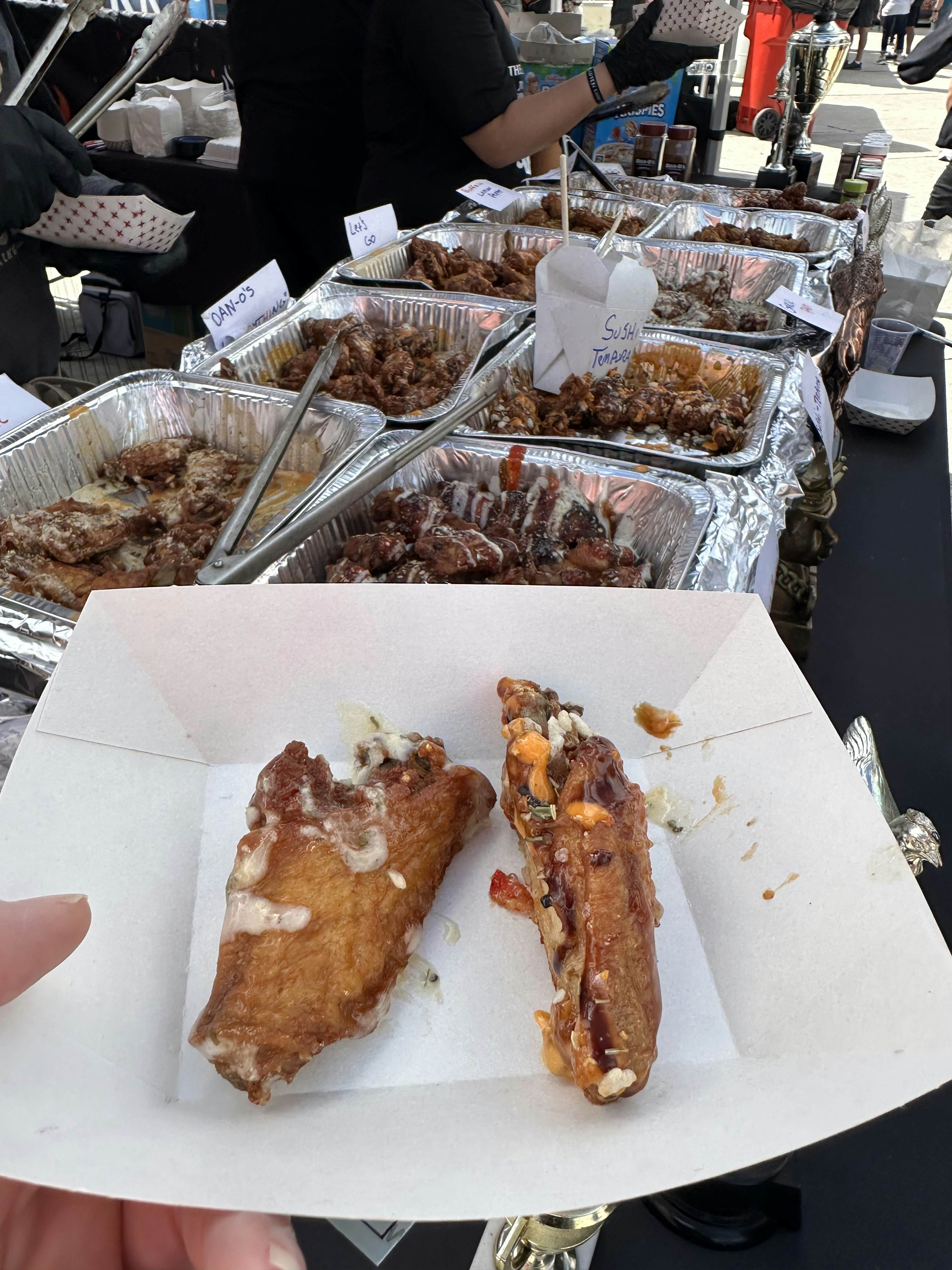 Aluminum trays of chicken wings in various sauces with tongs ready to be served