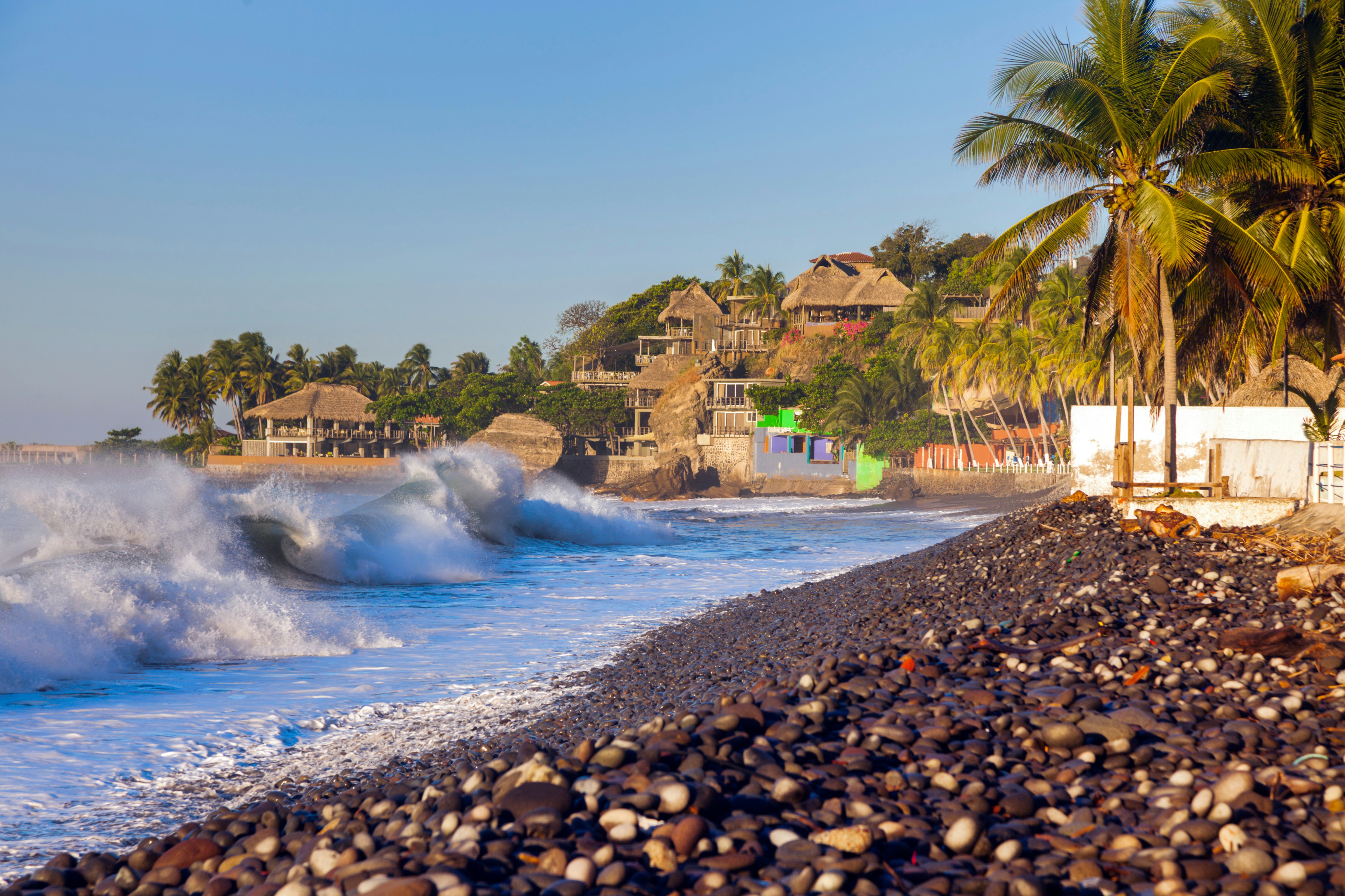 Waves break on a beach lined with pebbles, homes and palm trees