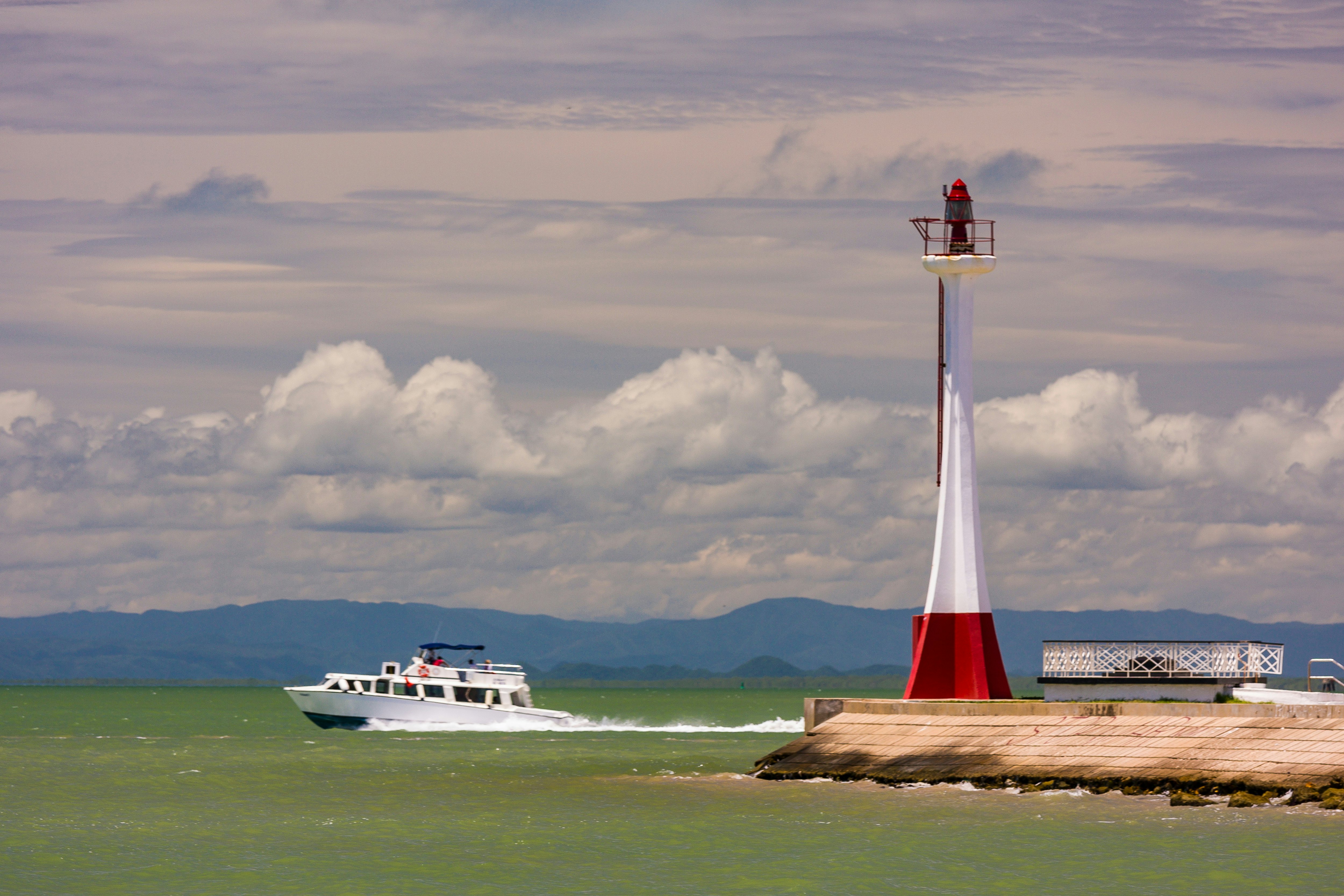 A power boat moves through greenish water past a narrow red-and-white lighthouse on a cloudy day.