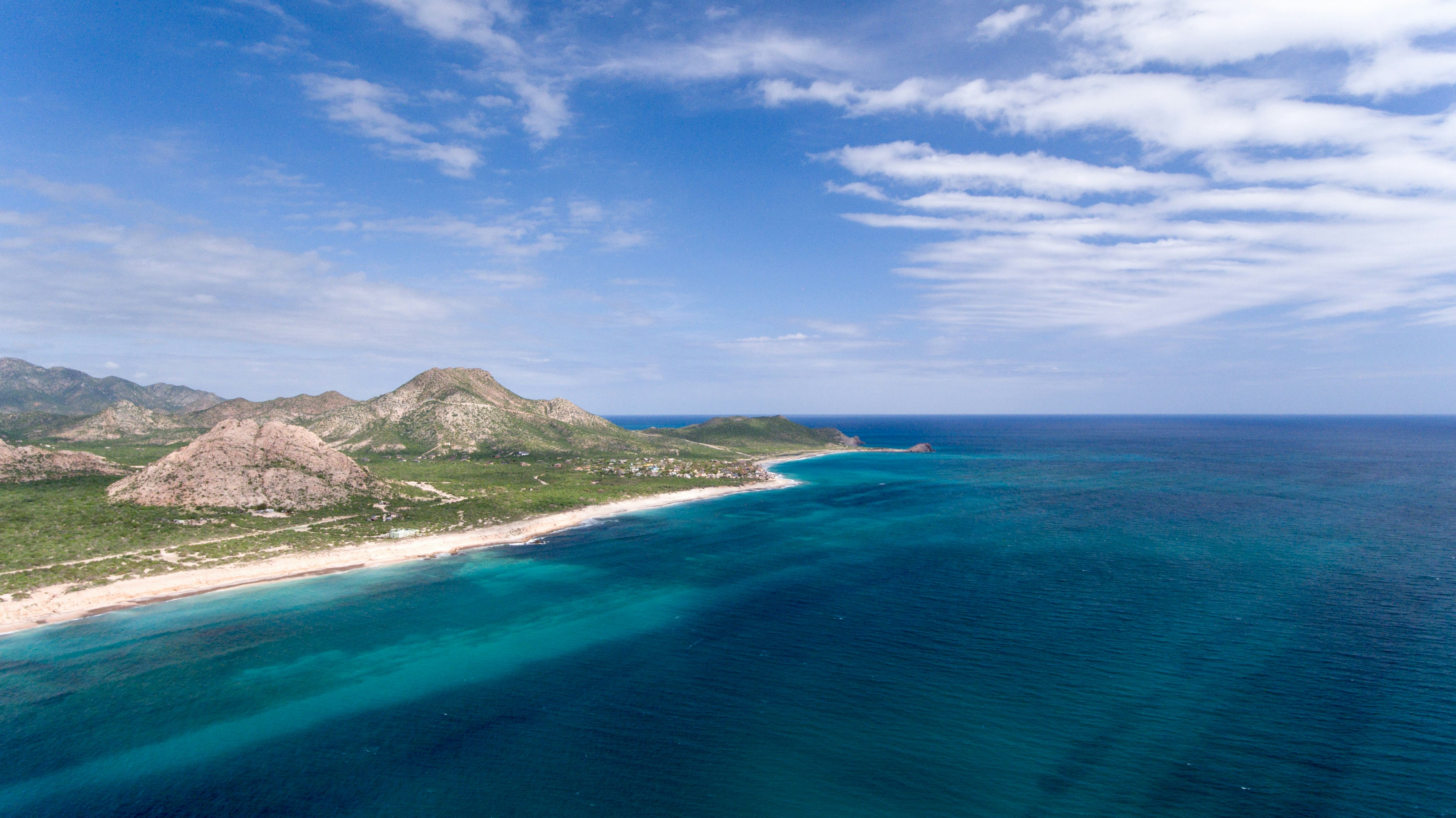 Aerial view of dark blue ocean with narrow sandy beach, grassland and rocky hills to the left on a sunny day.