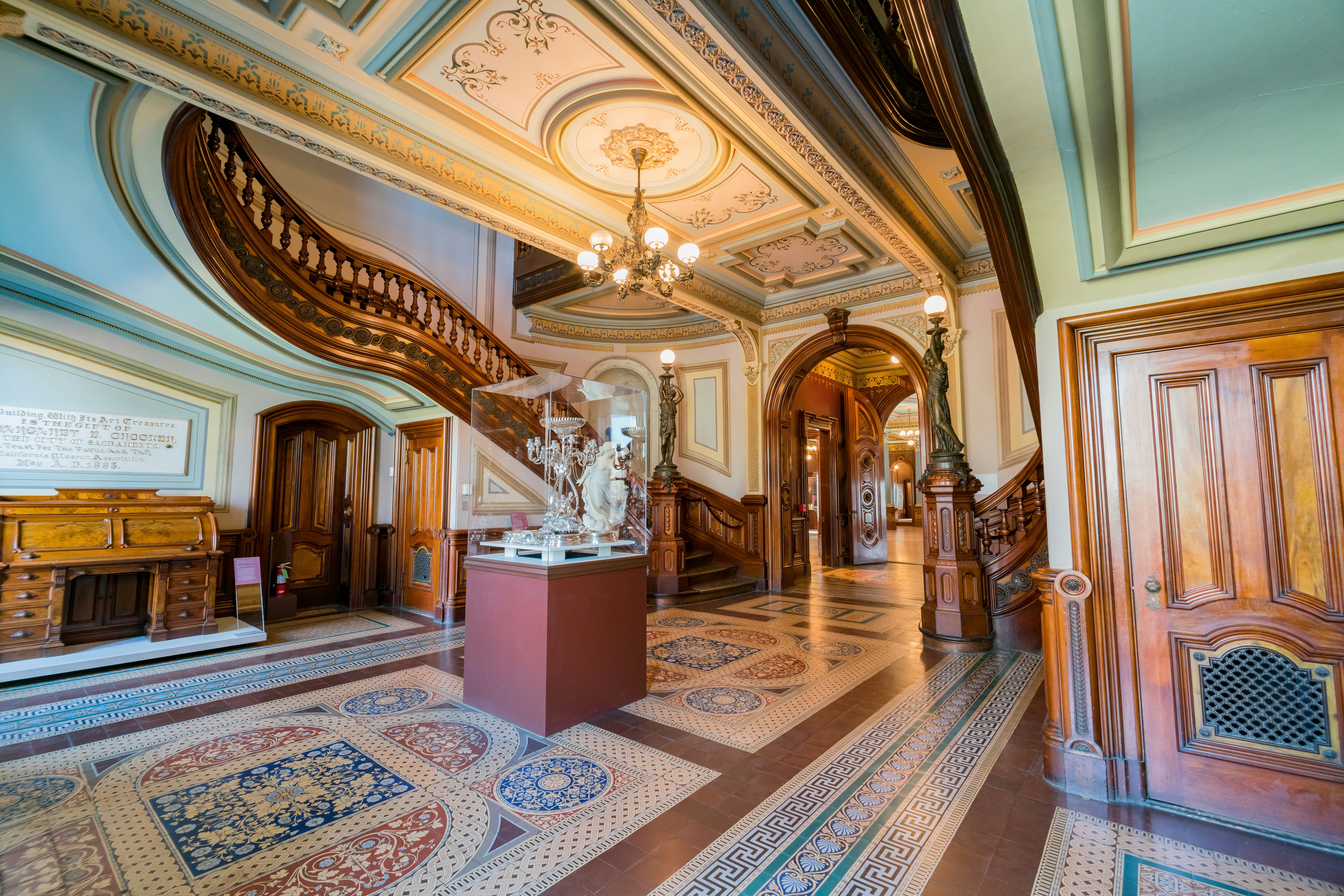 An entrance hall with intricately tiled floor, a decorative ceiling with chandelier and elaborate bannister on its curved staircase.