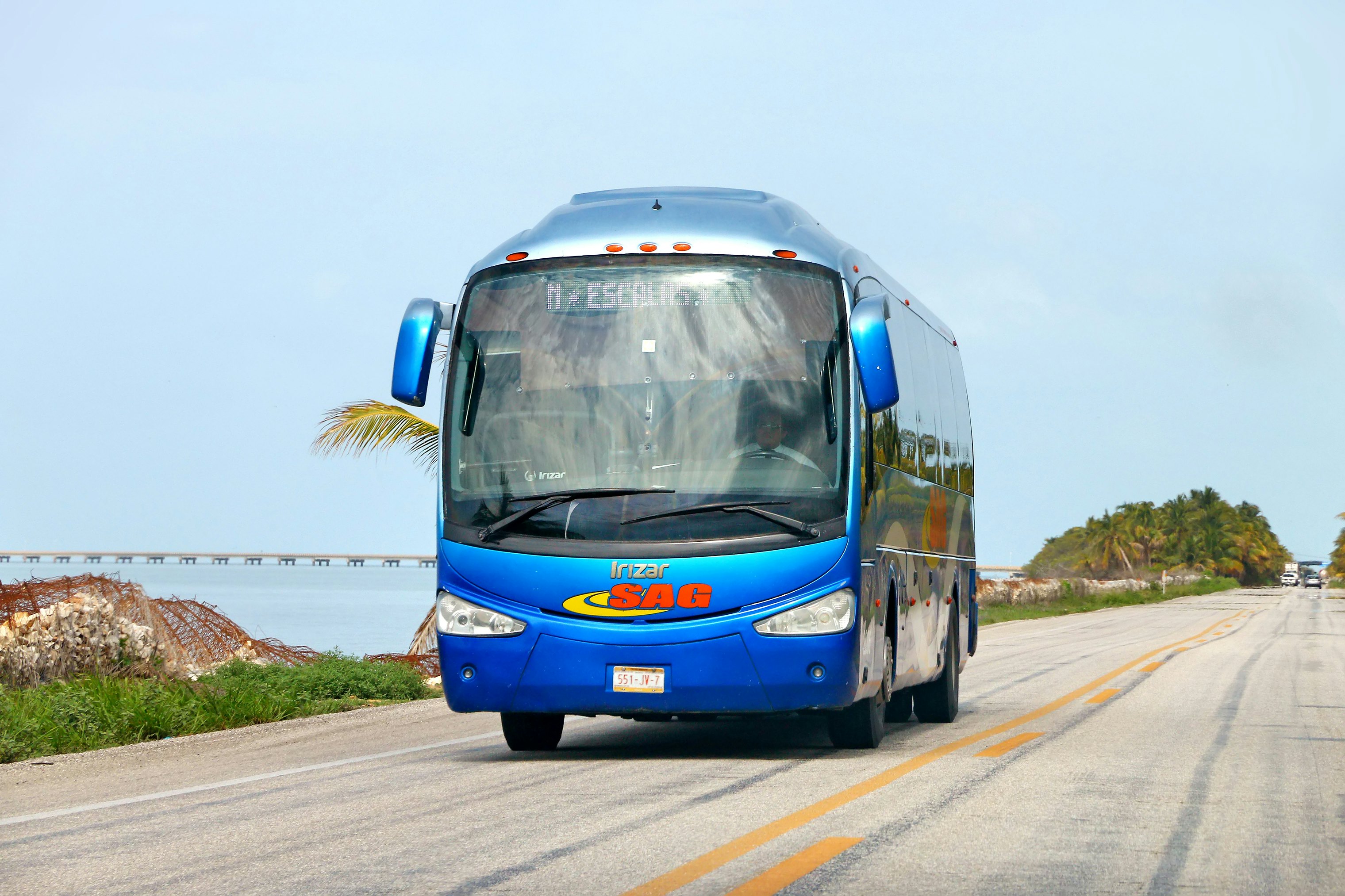 A blue-painted passenger coach drives on a highway next to a body or water.