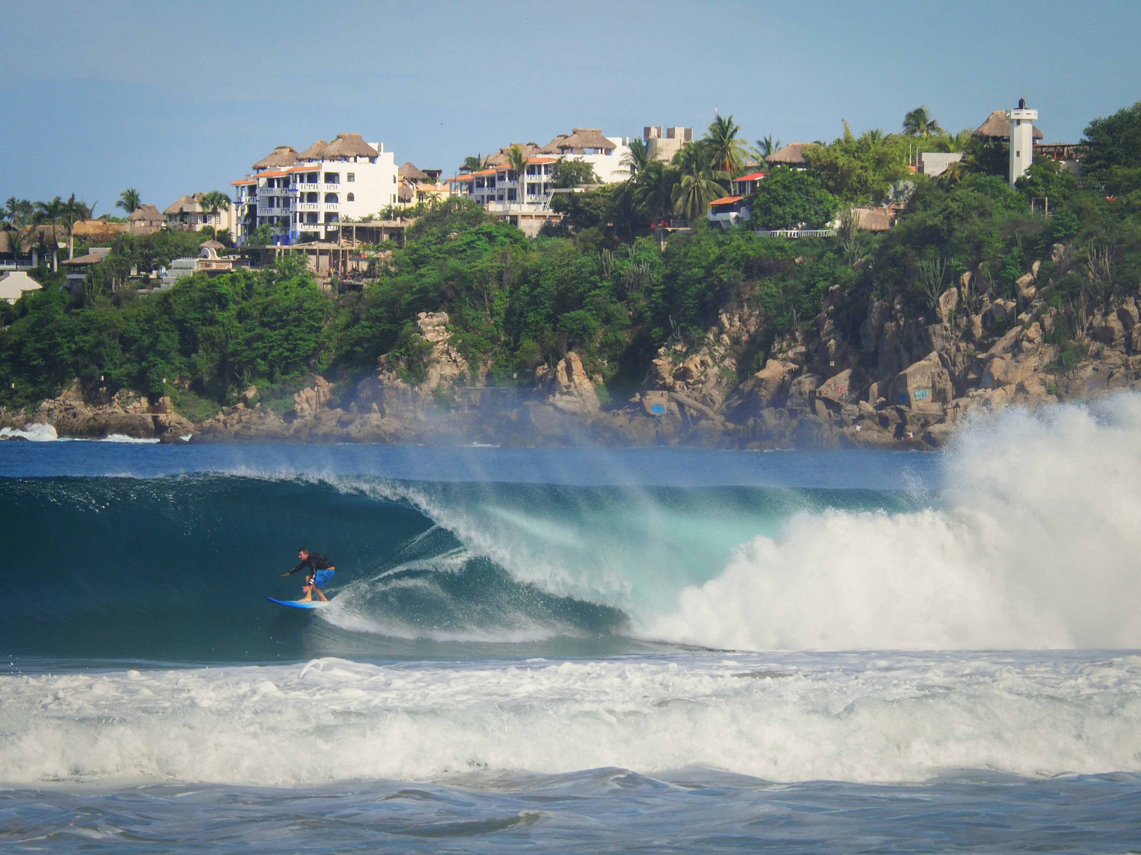 A surfer riding a large ocean wave with a hilltop town in the background on a sunny day.