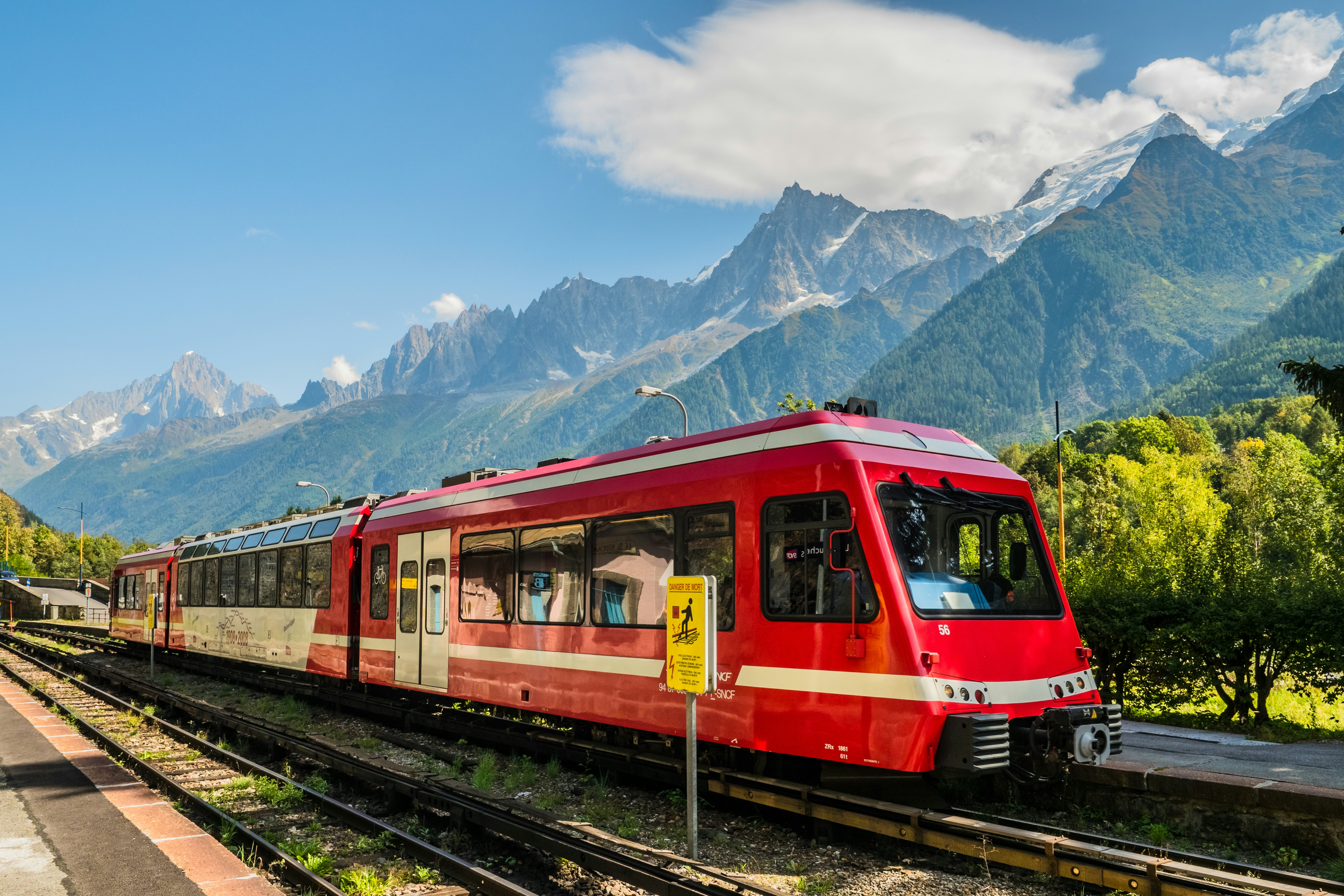 A train with three red cars travels on a track in a mountain valley with peaks rising in the distance.