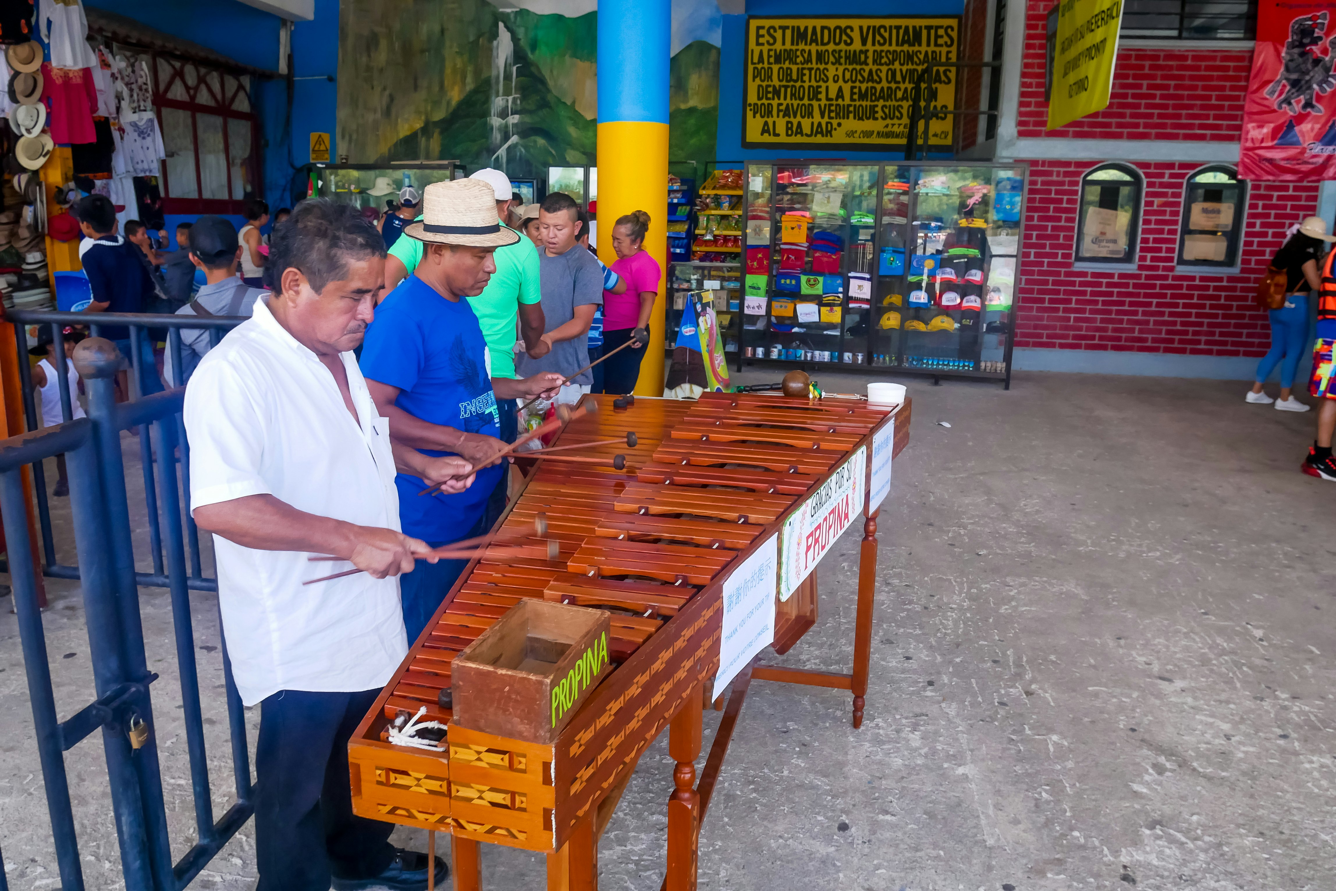 Two musicians play the marimba in an outdoor commercial area while other people shop and gather in the background.