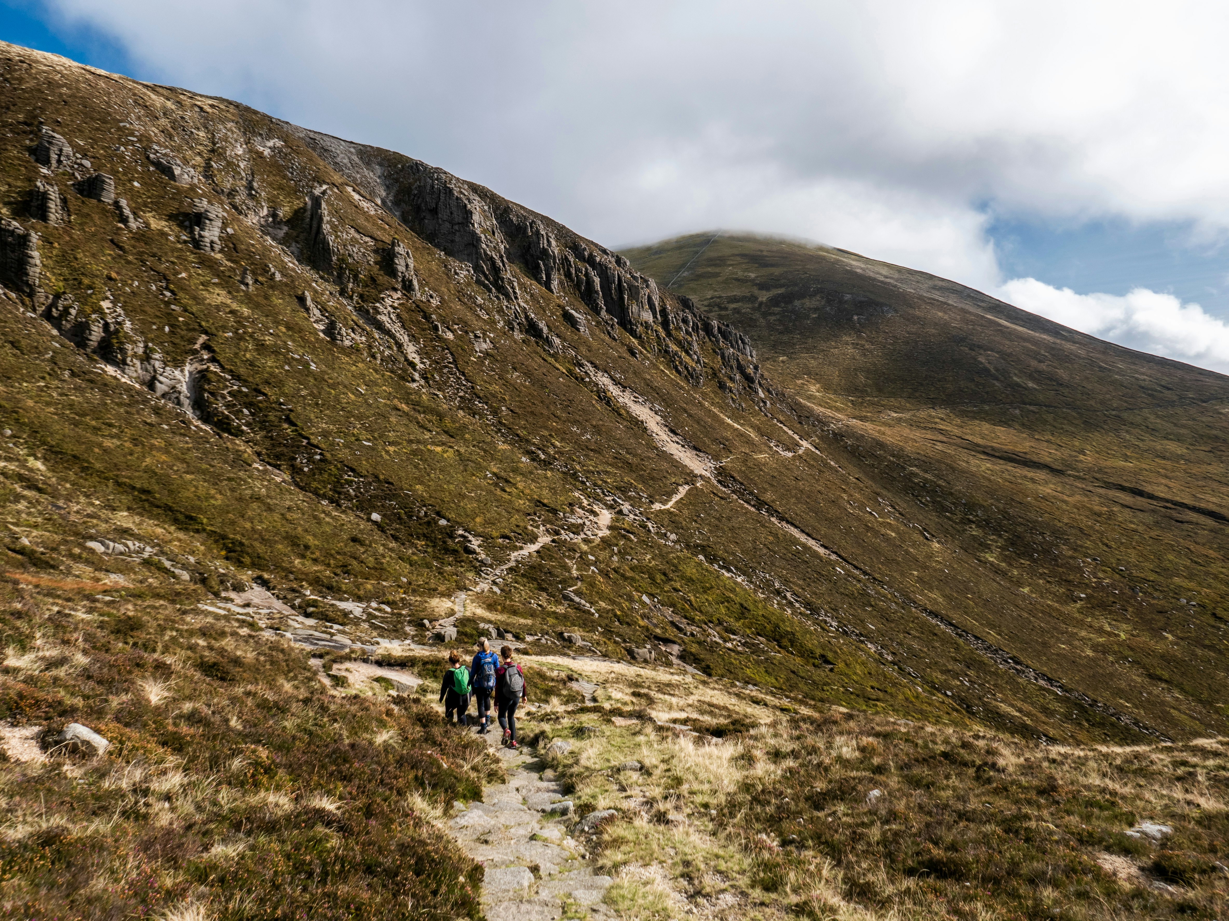 A wide shot of hikers following a trail along a ridge of mountains covered in shrubs.