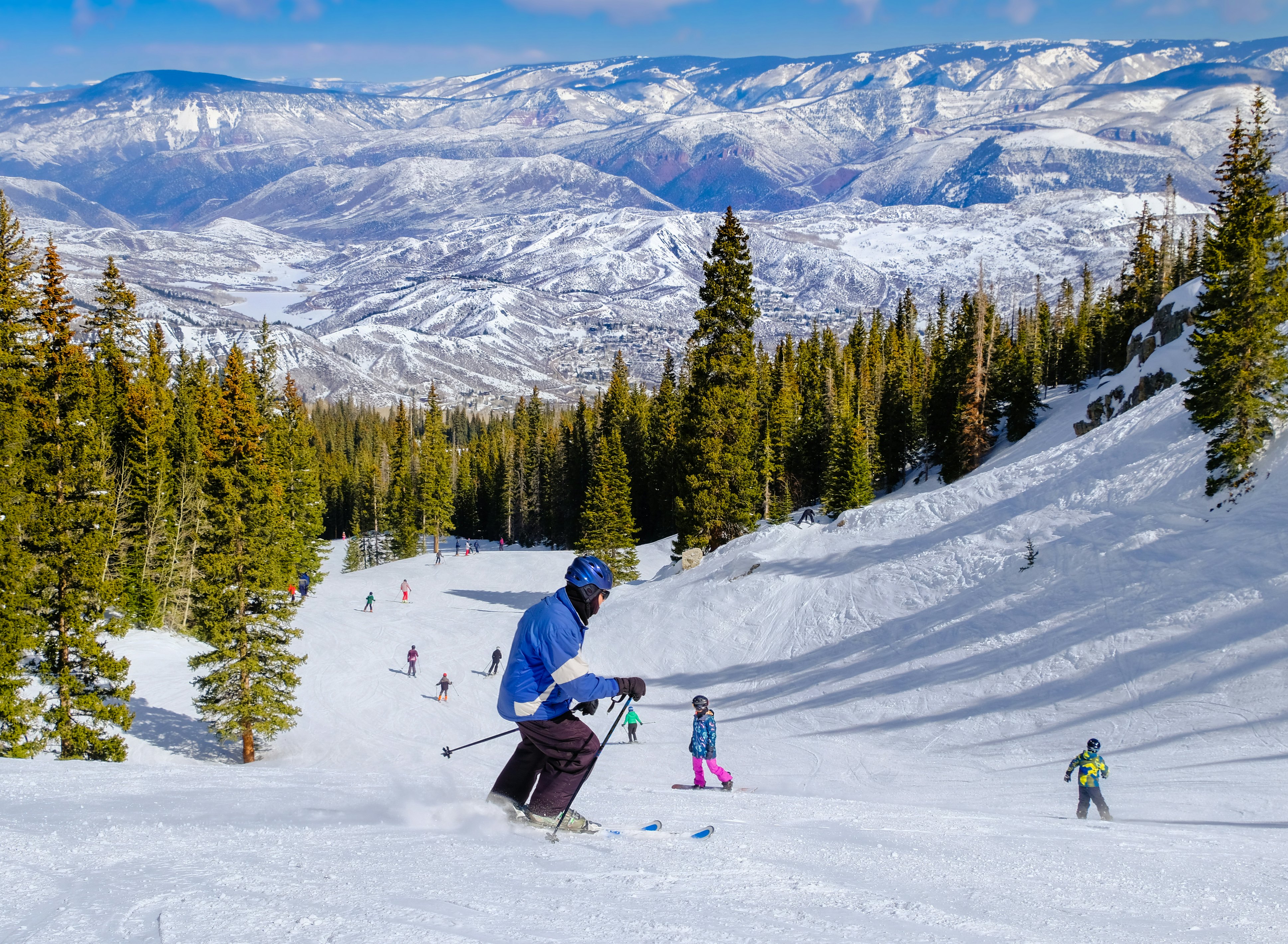 A skier going downhill toward other skiers, with views of the mountains and alpine trees.