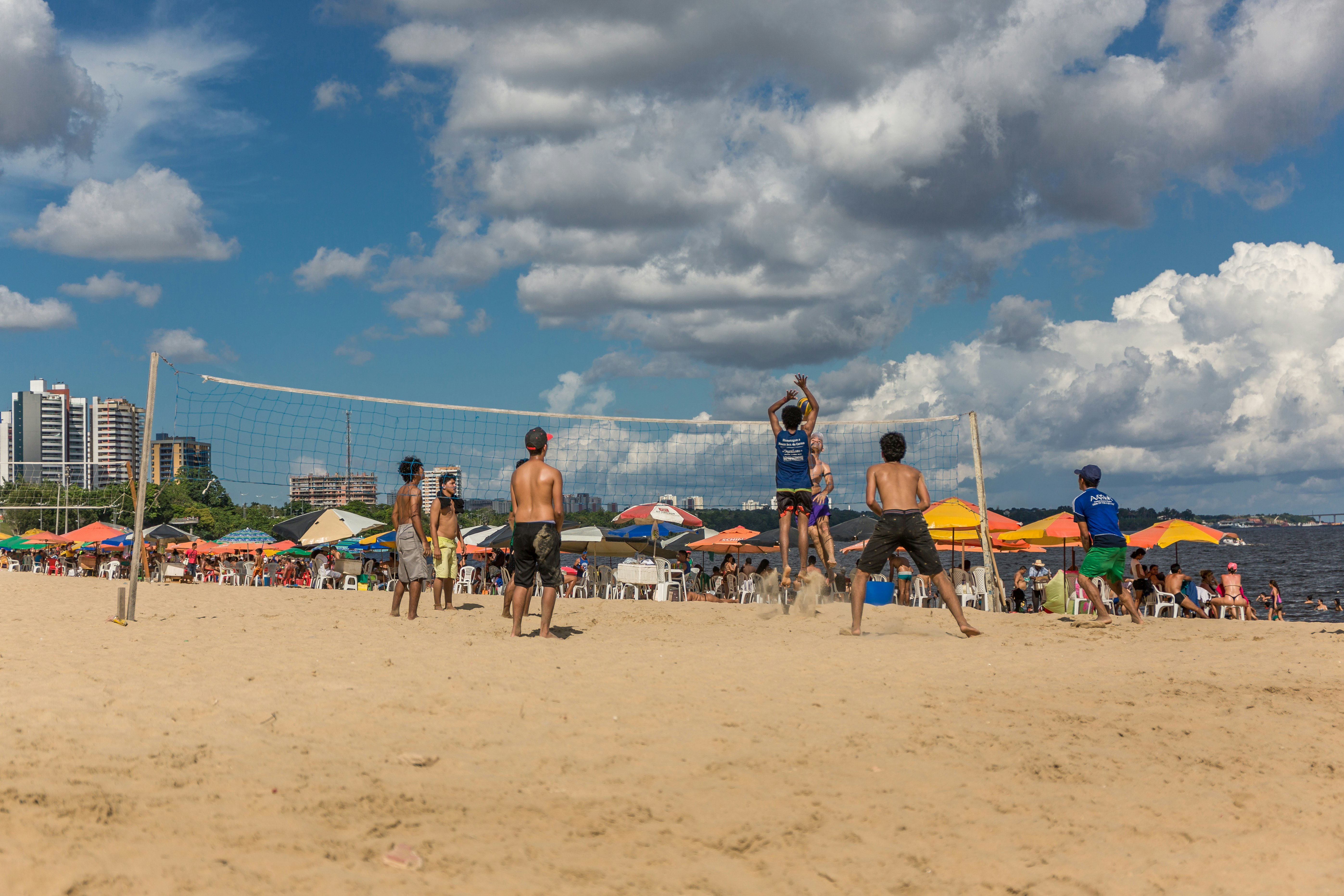 Group of local Brazilians play beach volleyball on the urban Ponta Negra beach in the Amazon while people relax sitting on chairs under sun umbrellas in the background