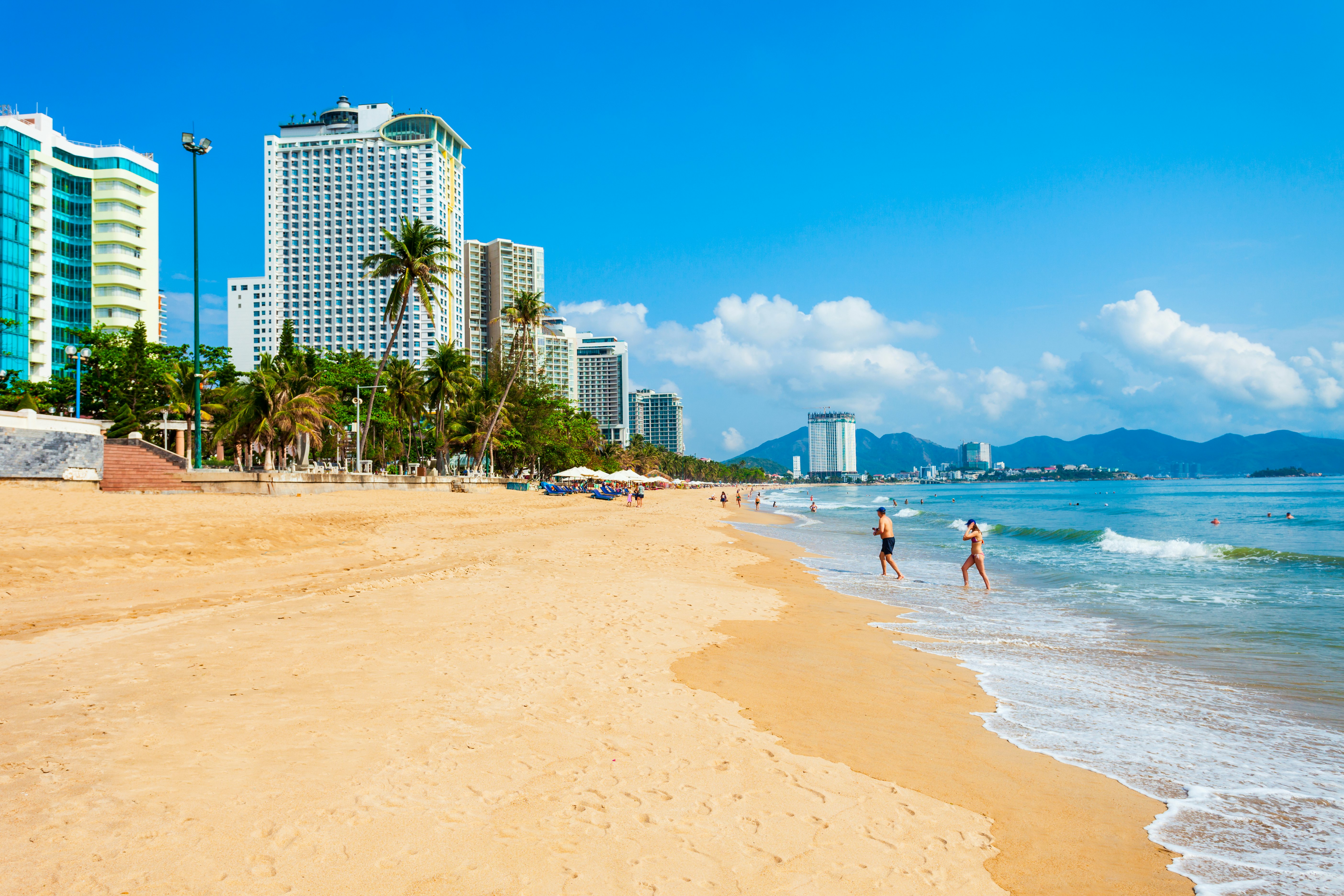 Two people wander out of the surf on a long golden stretch of sand next to a high-rise city skyline.