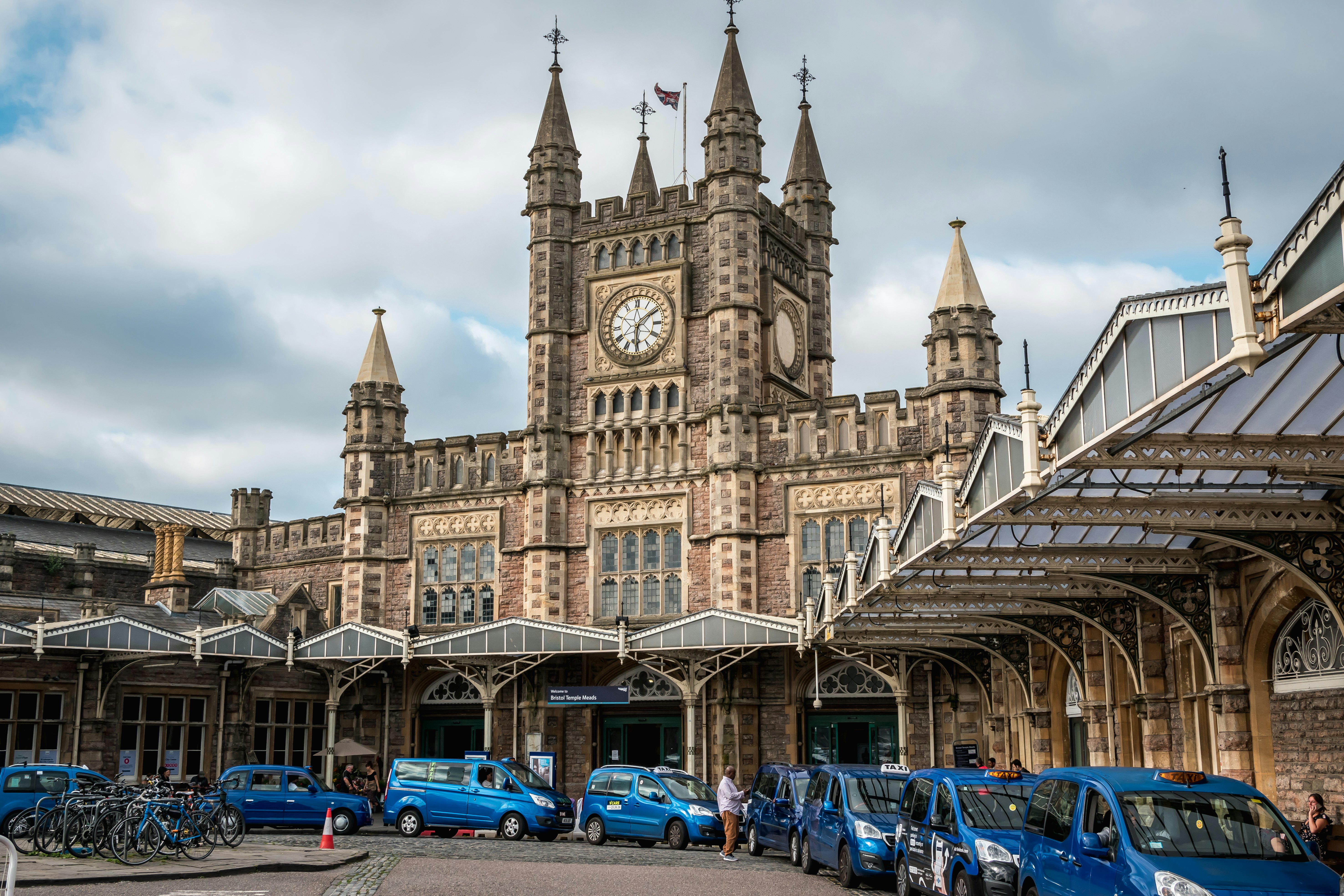 Blue taxis line up outside the grand facade of a train station with a central Gothic clock tower.