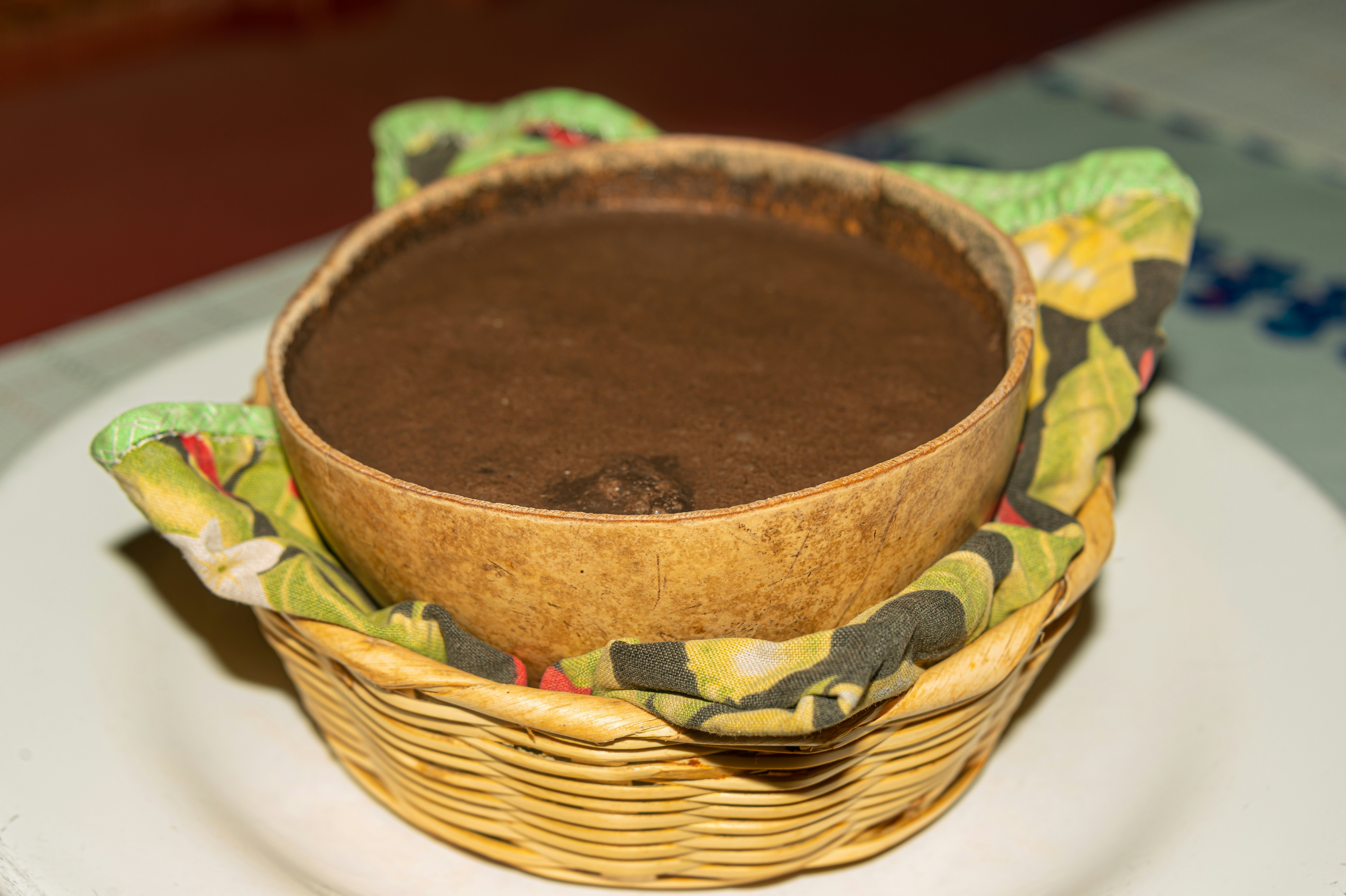 Pozol traditional drink (chocolate brown-colored thick liquid) in a large round bowl tucked into a woven basket with a printed texile between the bowl and basket.