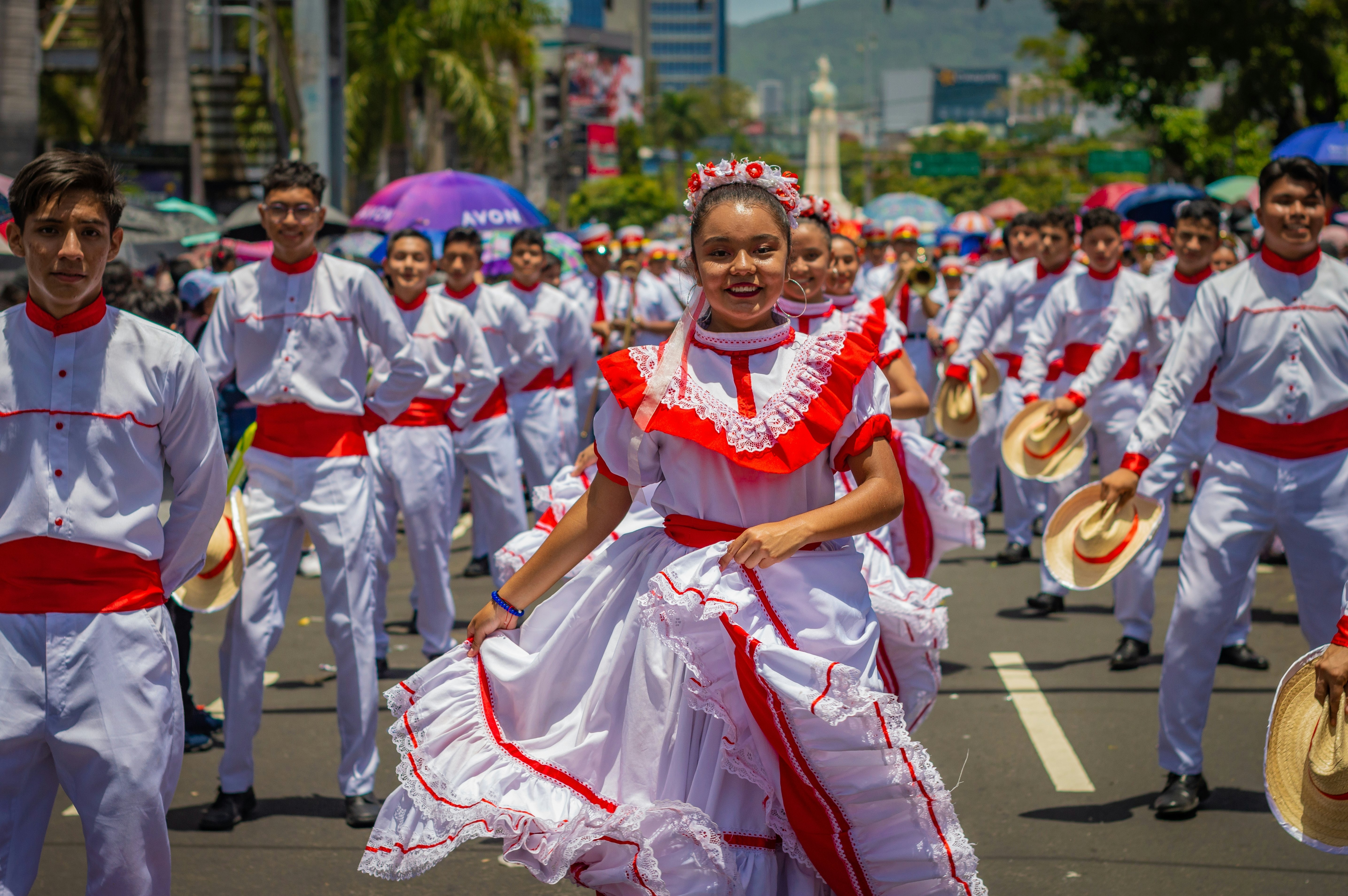 A street parade with smiling dancers and performers wearing white clothing with red trimmings. The men all carry a straw hat.