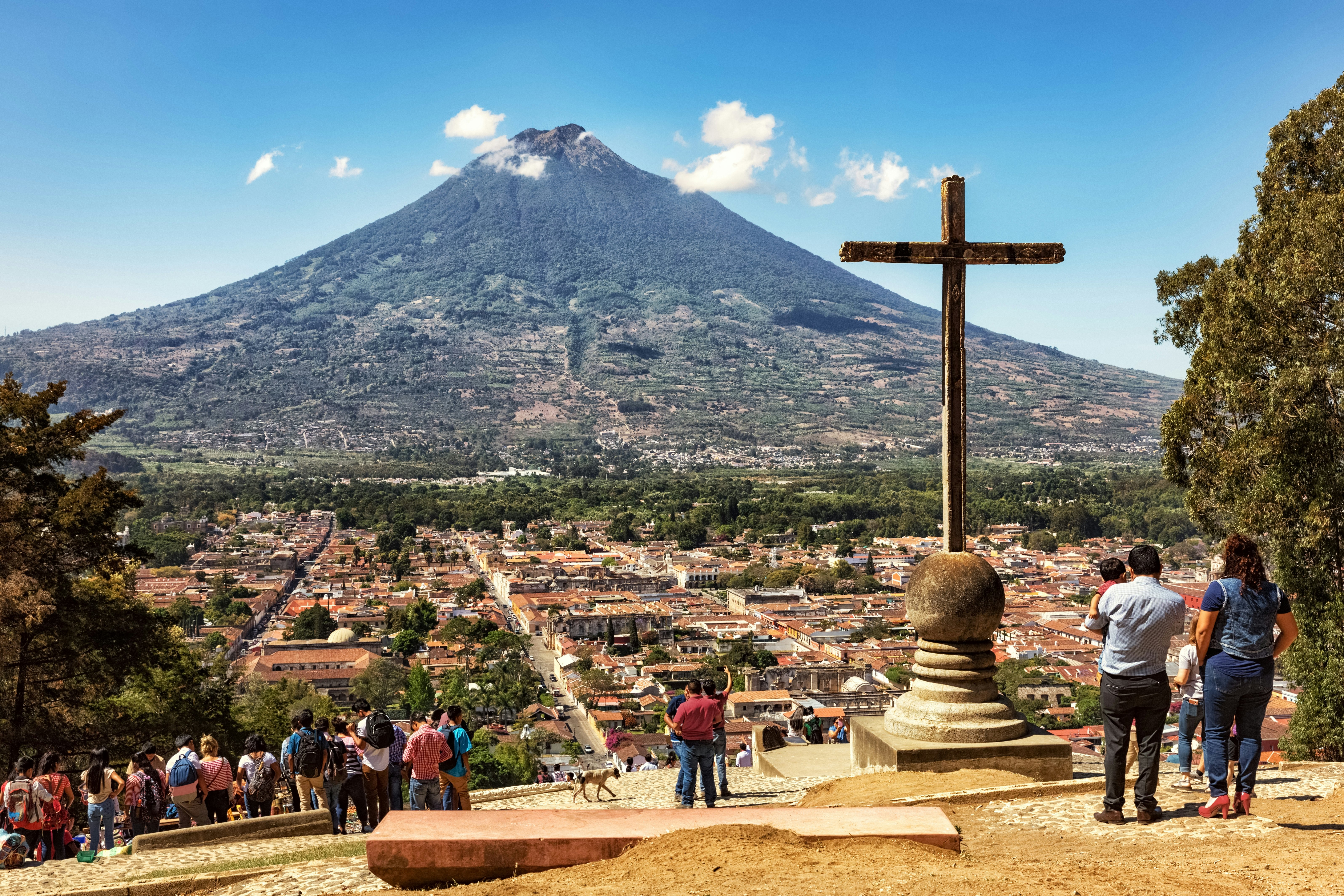 People stand on a hillside with a large cross overlooking a city of red-roofed buildings and a volcano in the distance on a sunny day.