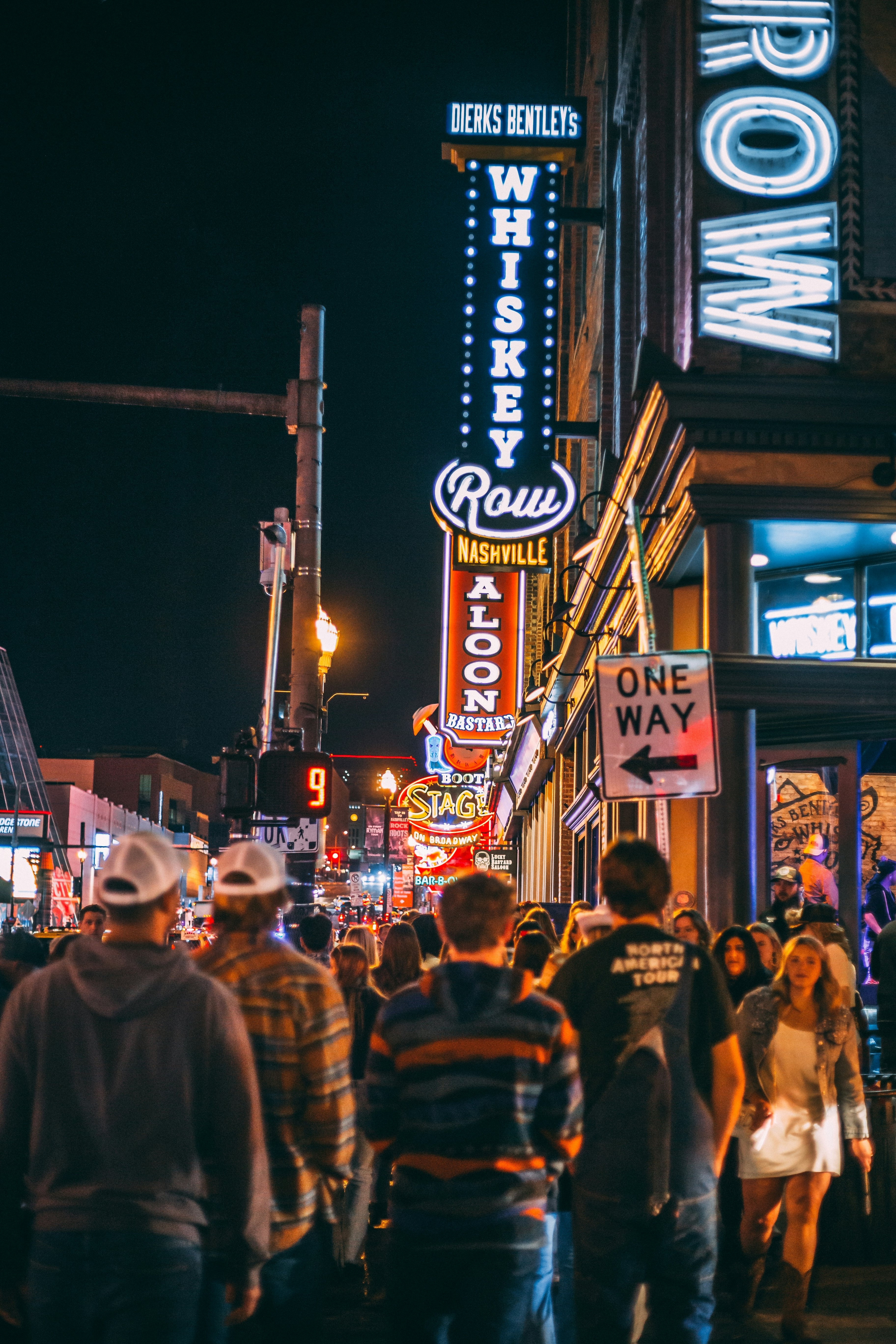 People walking on a crowded city street at night with neon signs reading "Whiskey Row" and "Nashville."