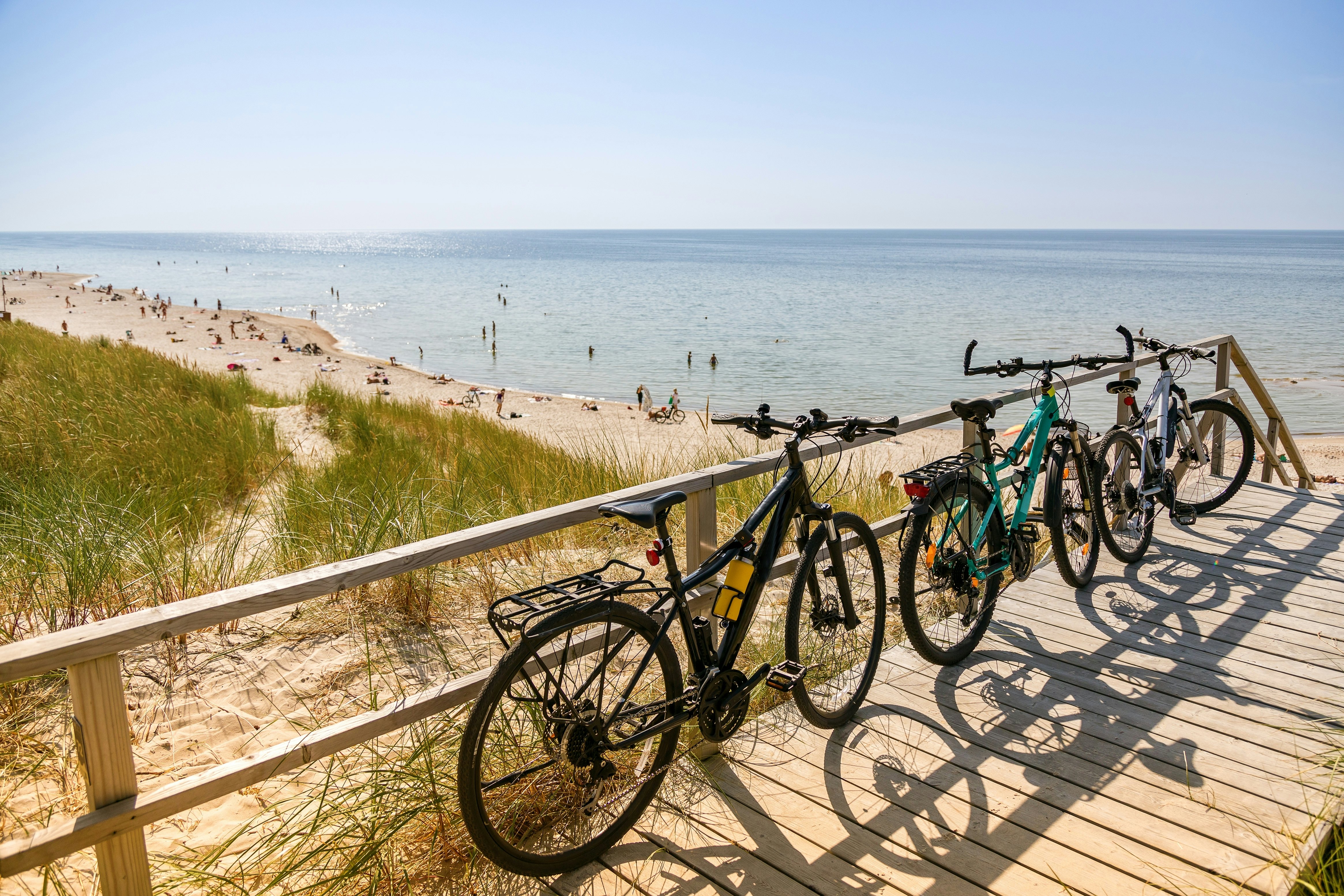 Three bikes lean on railings along a boardwalk above a beach backed by grassy sand dunes in a summer's day. People paddle and sunbathe on the beach below.