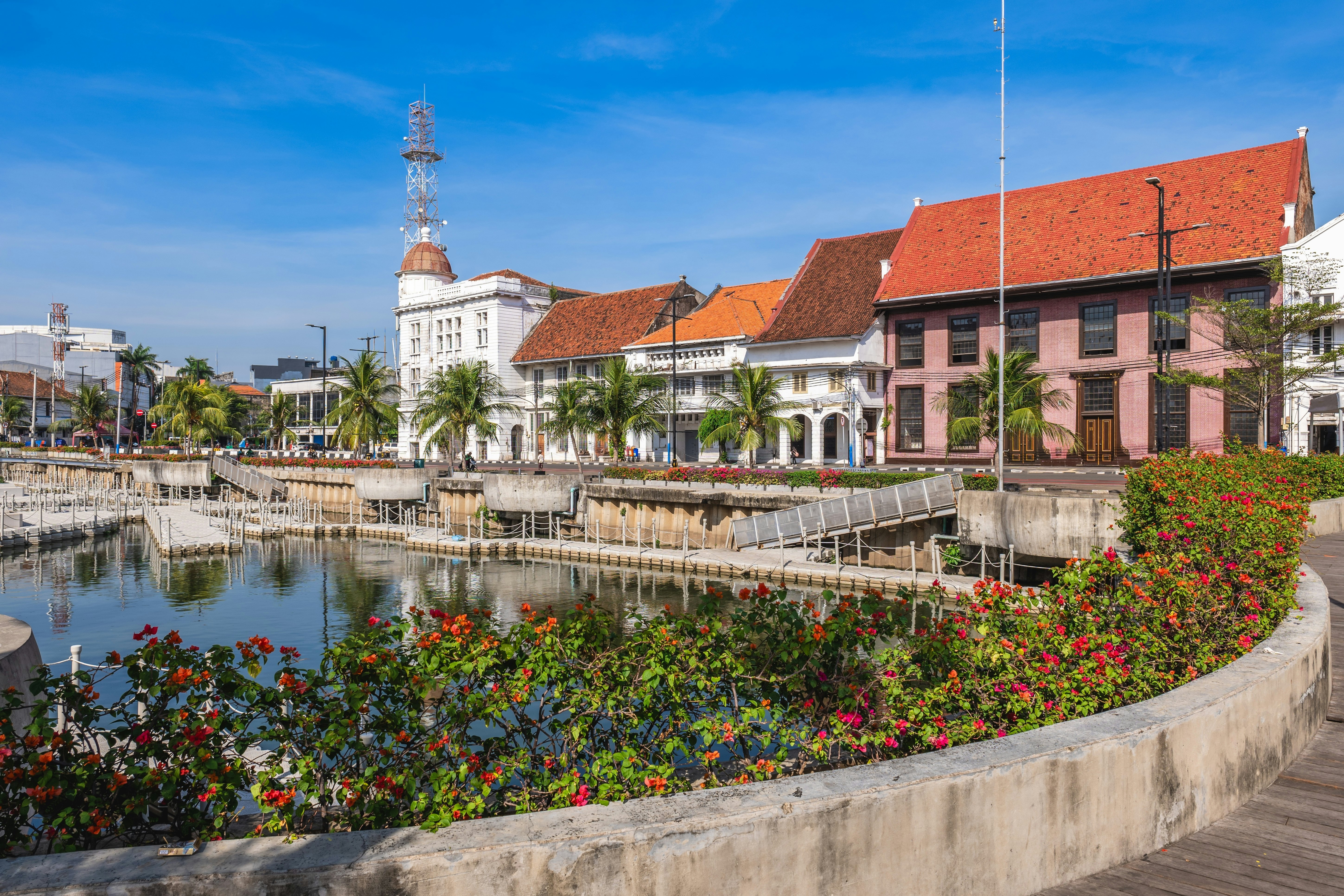 Small buildings on a tree-lined street with a flower-lined body of water nearby in Jakarta's Old Town.