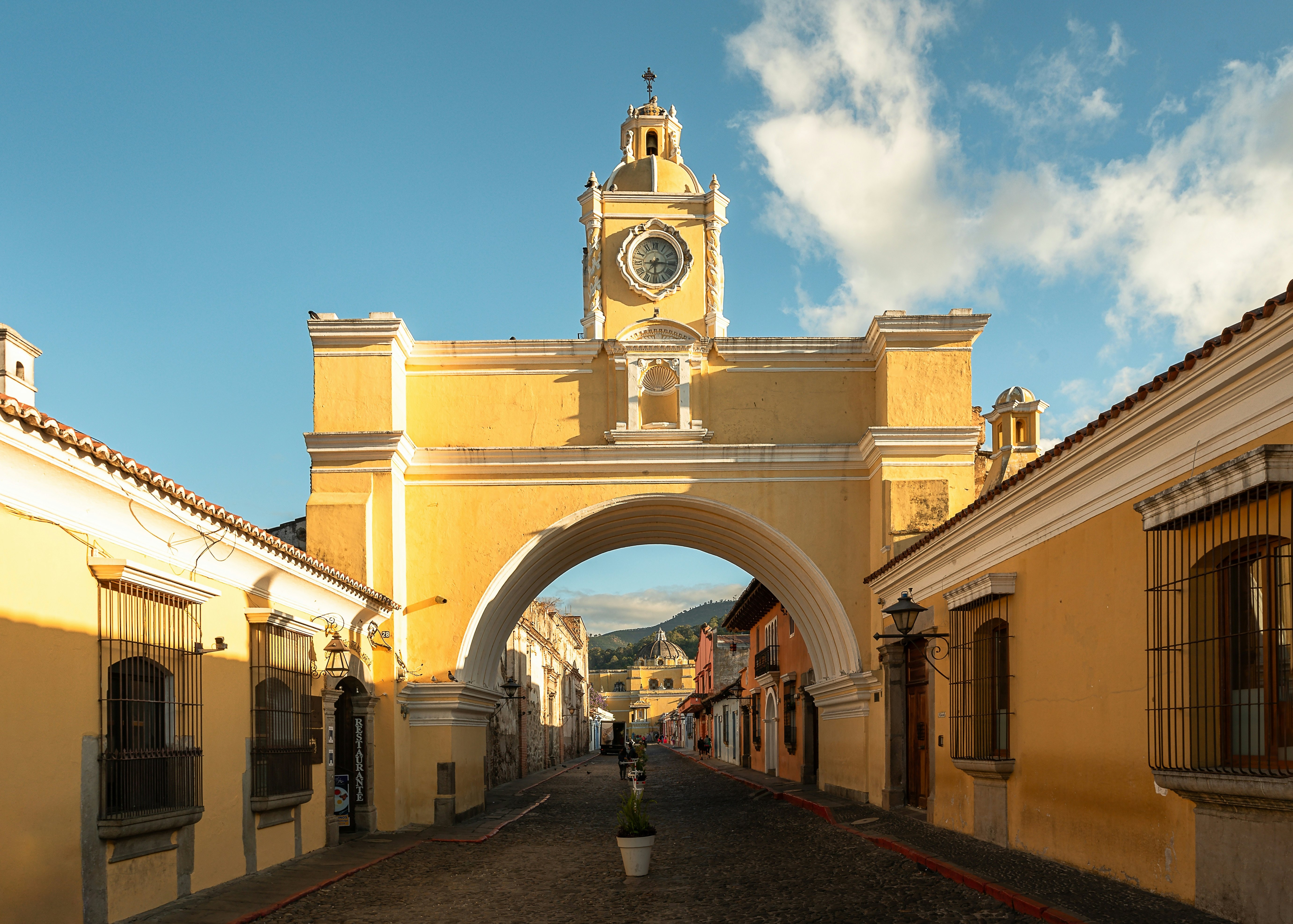 A large yellow archway with a clock on top over a cobblestone street on a city day.