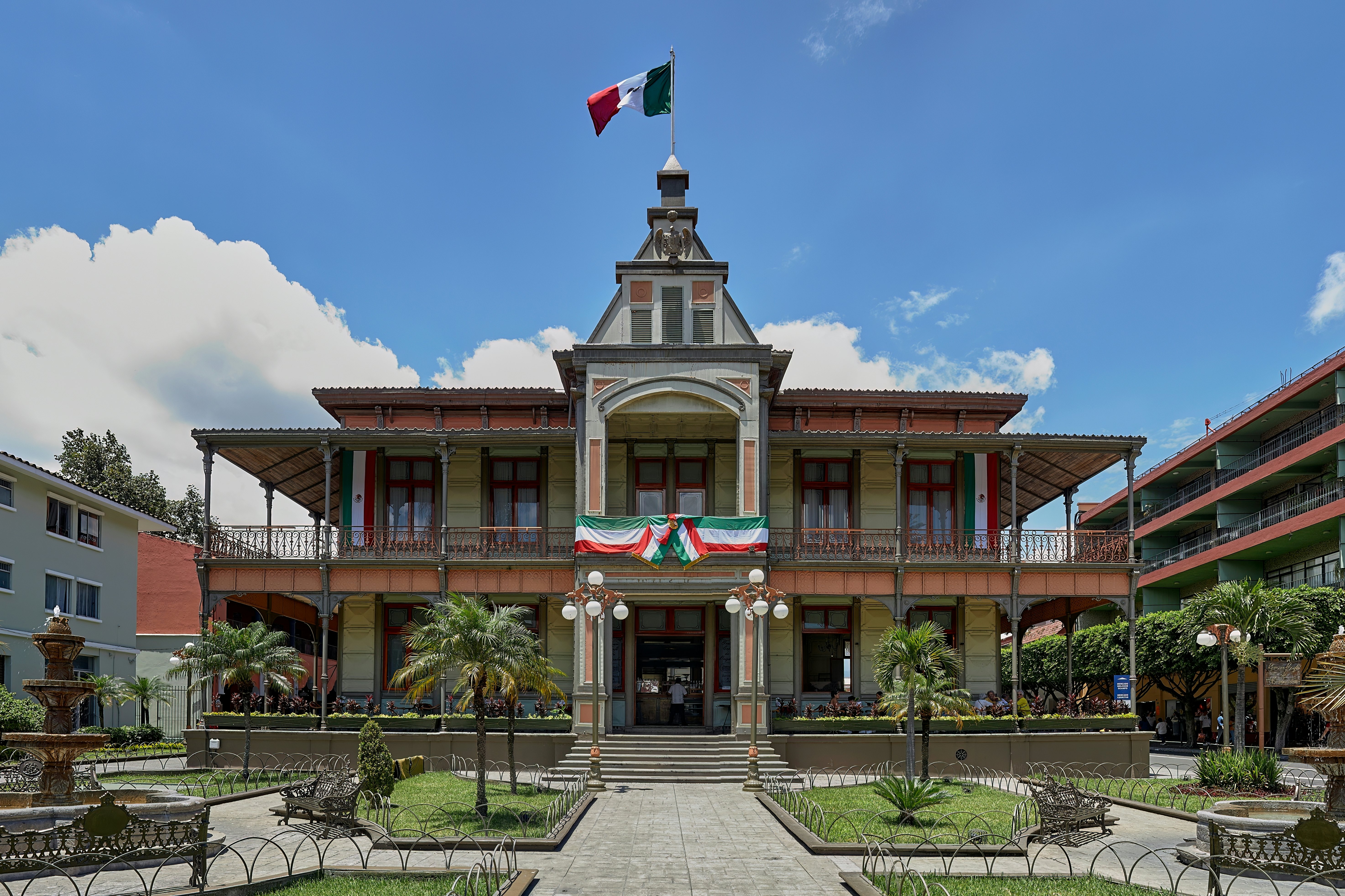 A historic building with a covered porch and balcony seen from a formal garden square on a sunny day.