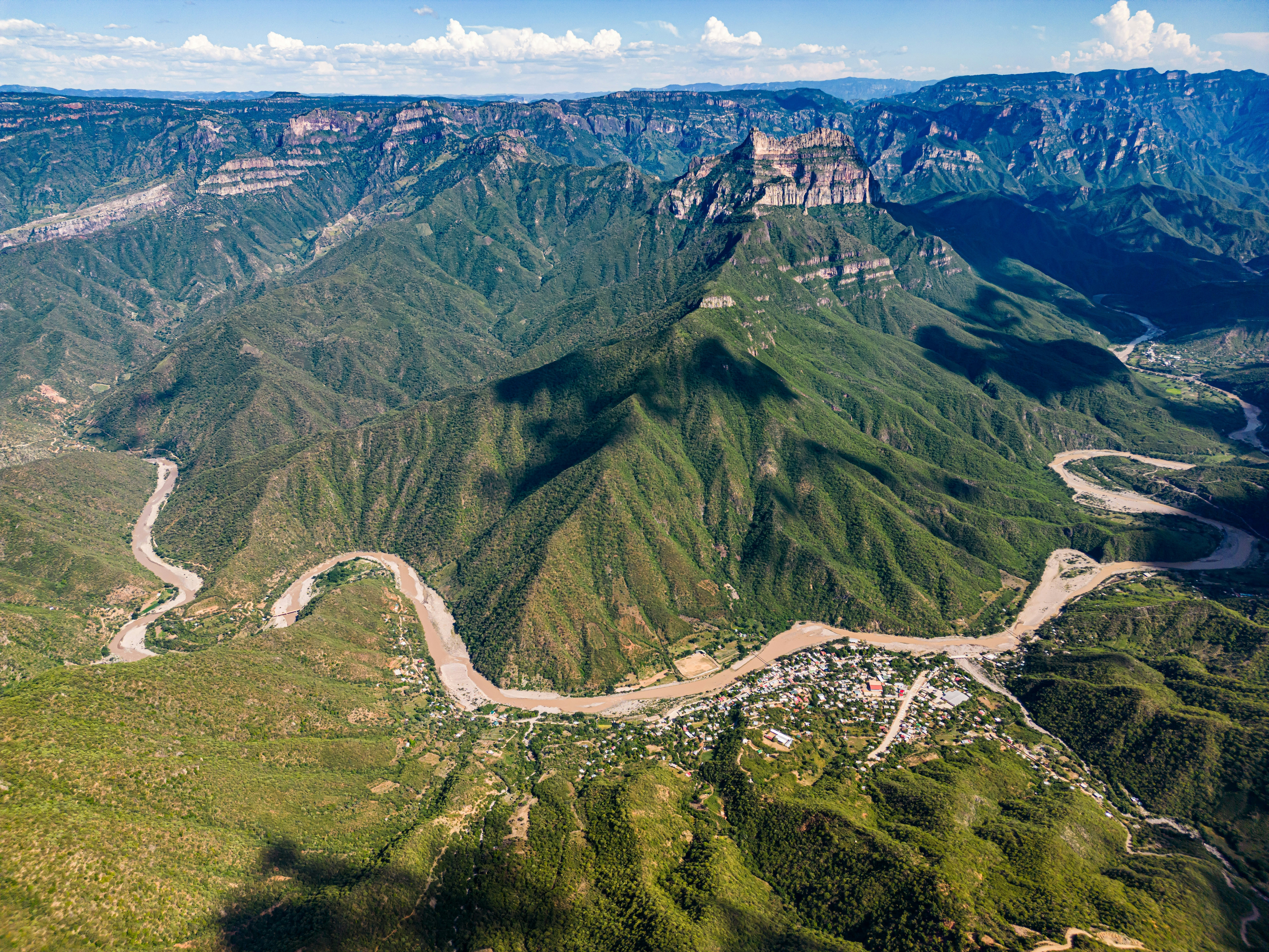 Aerial view of winding road going through dramatic canyon between forest-covered peaks on a sunny day.