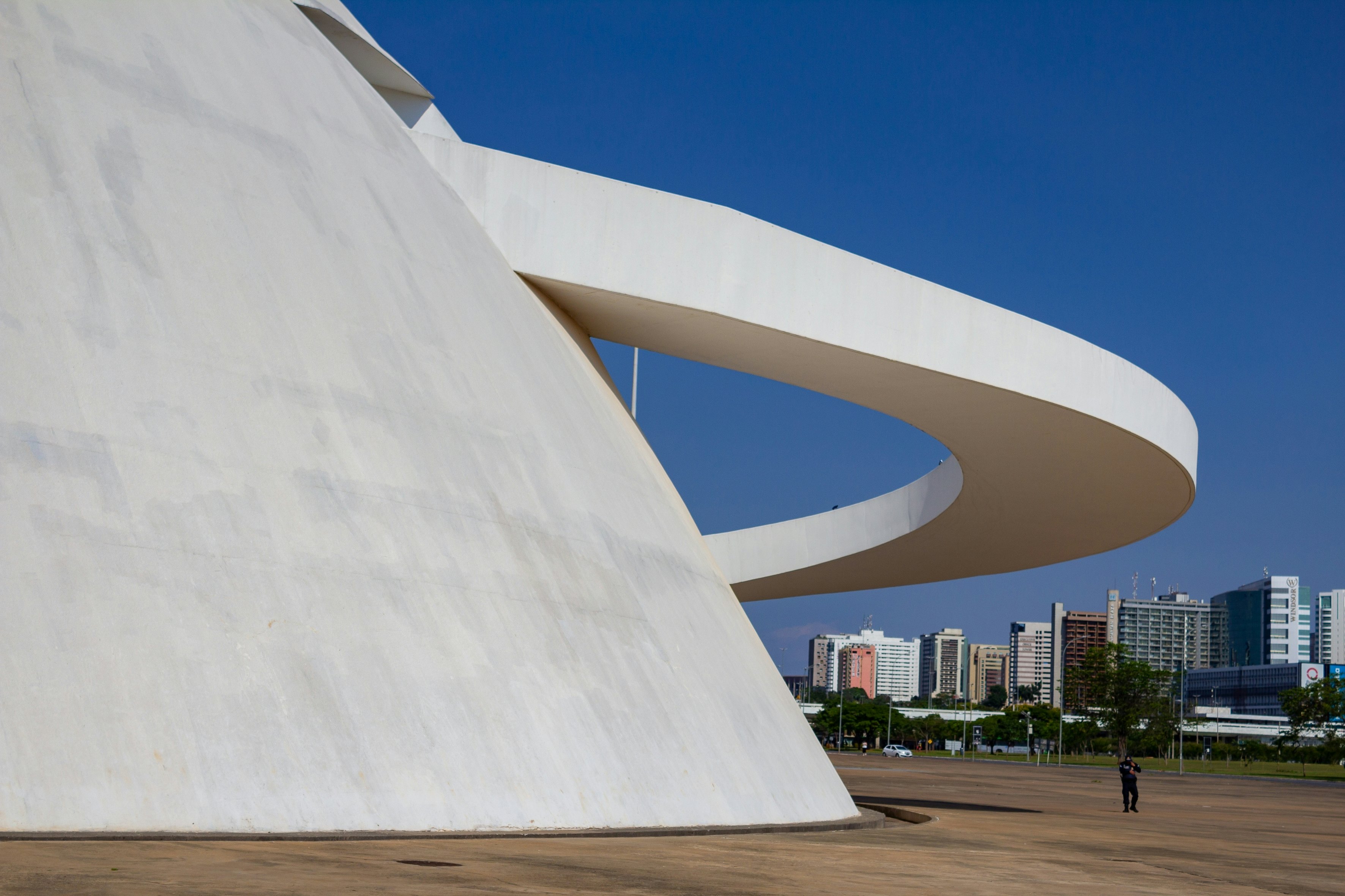 A single person stands under a curved white element extending out around a round white building under clear blue sky in Brazil.