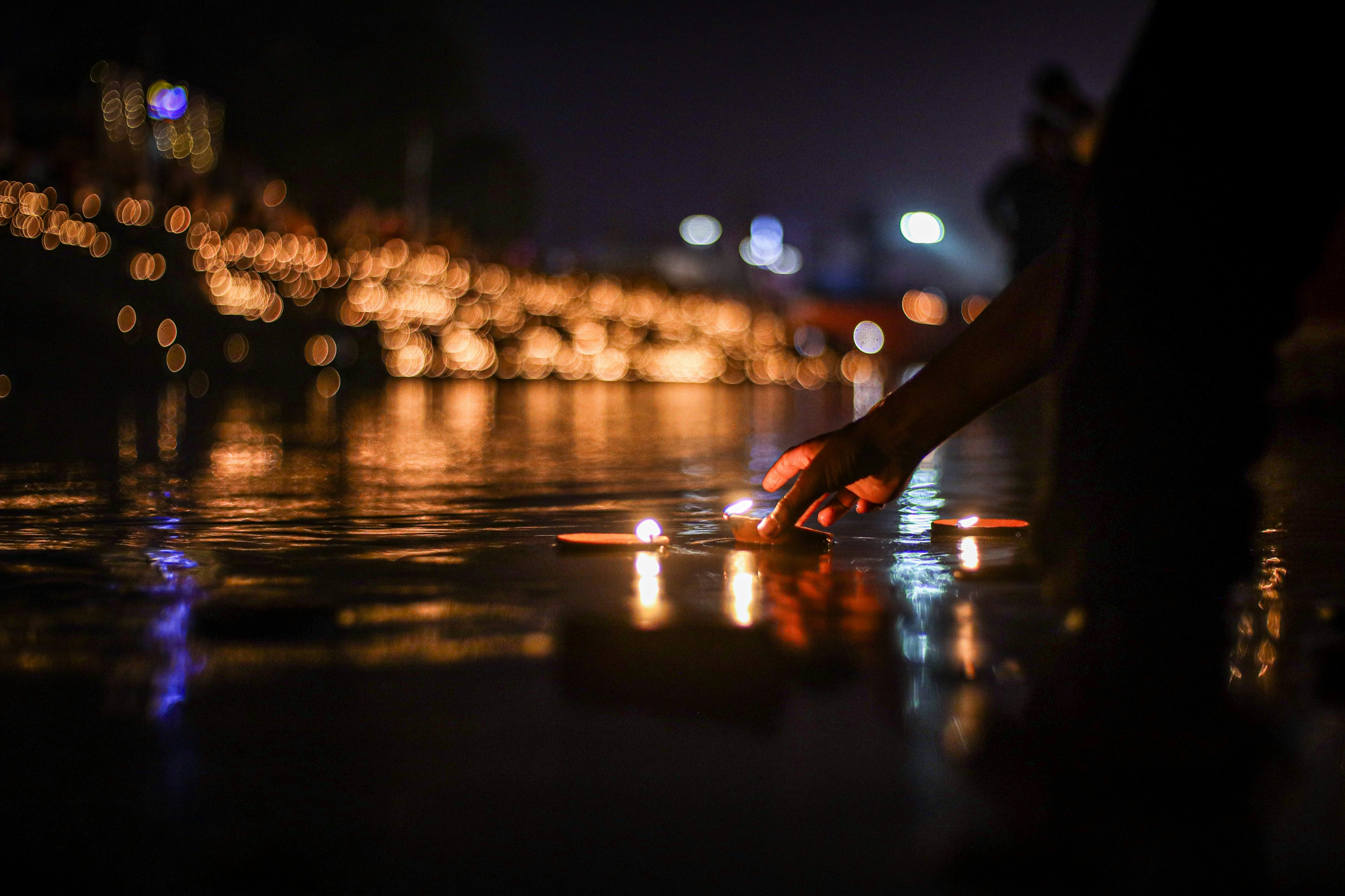 A person set a lamp with a flame on the water at night. Glowing lights of a city are seen in the distance.