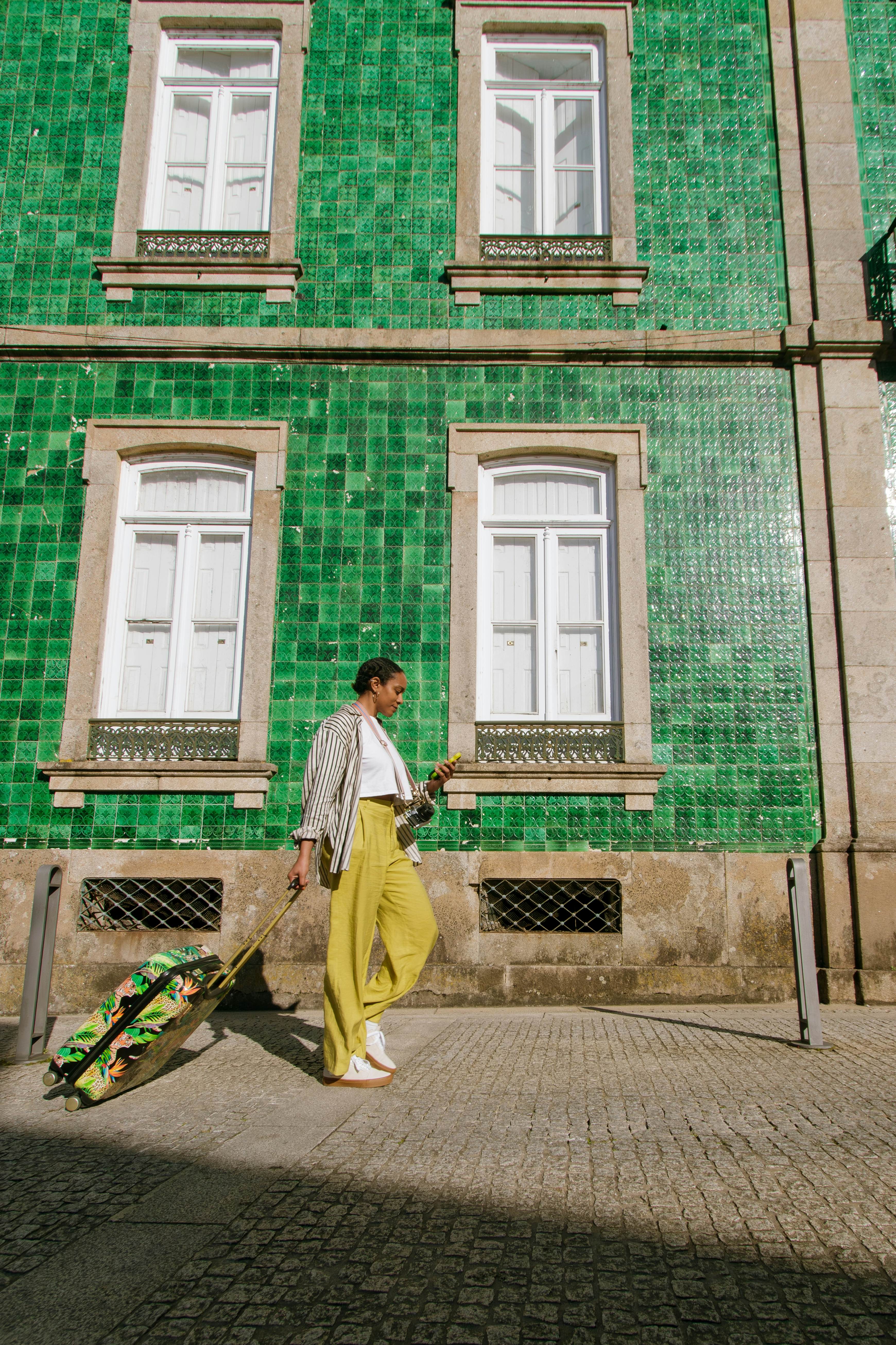 Vertical shot of black female tourist with suitcase walking in front a tiled green historical building checking her phone for directions on a sunny day carrying her luggage , License Type: media, Download Time: 2024-09-25T22:16:04.000Z, User: Norma.PrauseBrewer_LonelyPlanet, Editorial: false, purchase_order: 56530, job: Global Publishing WIP, client: Portugal 14, other: Norma Brewer