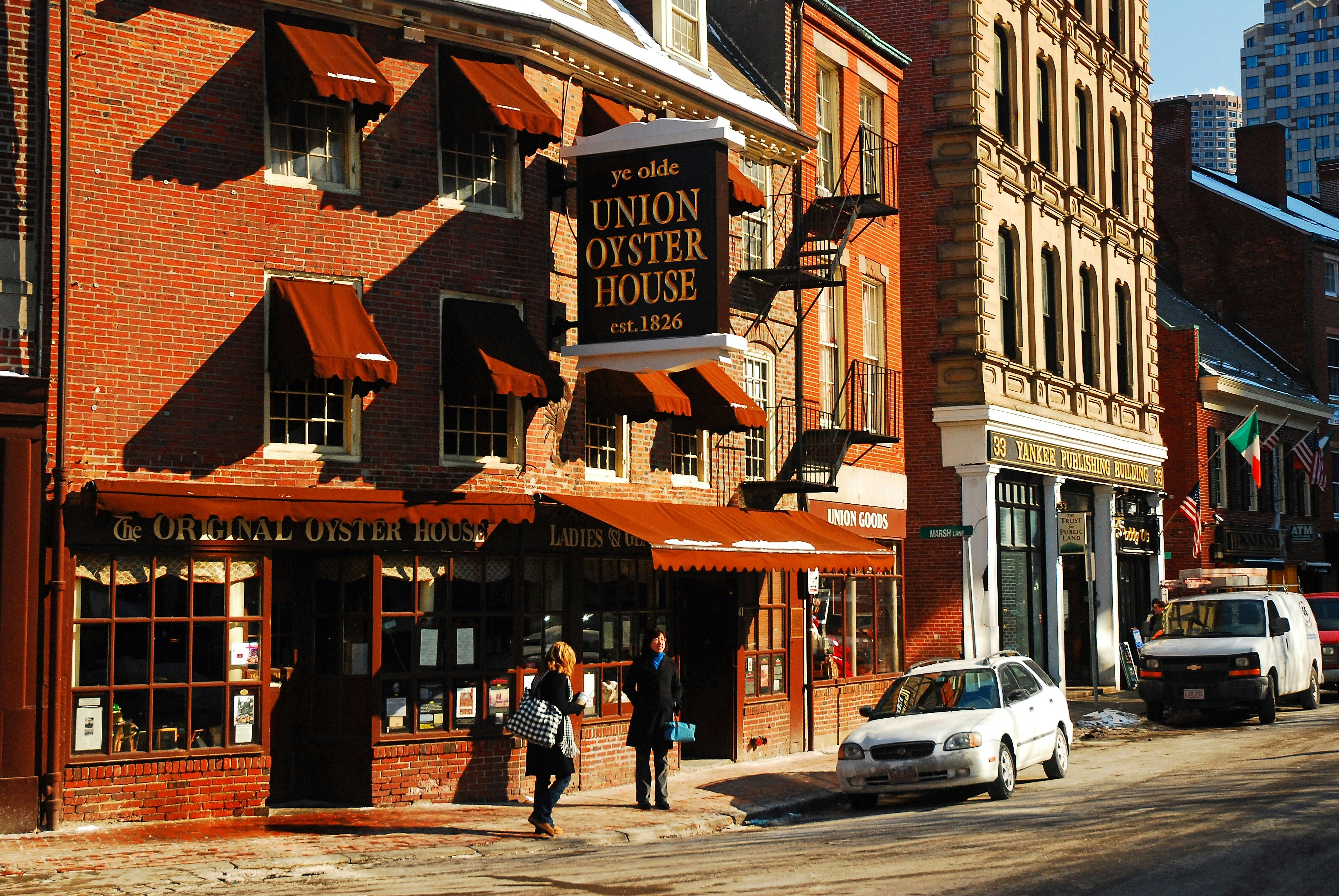 Two people walk by a red-brick restaurant with a large sign that says "Ye Olde Union Oyster House, est. 1826" on a sunny winter's day.
