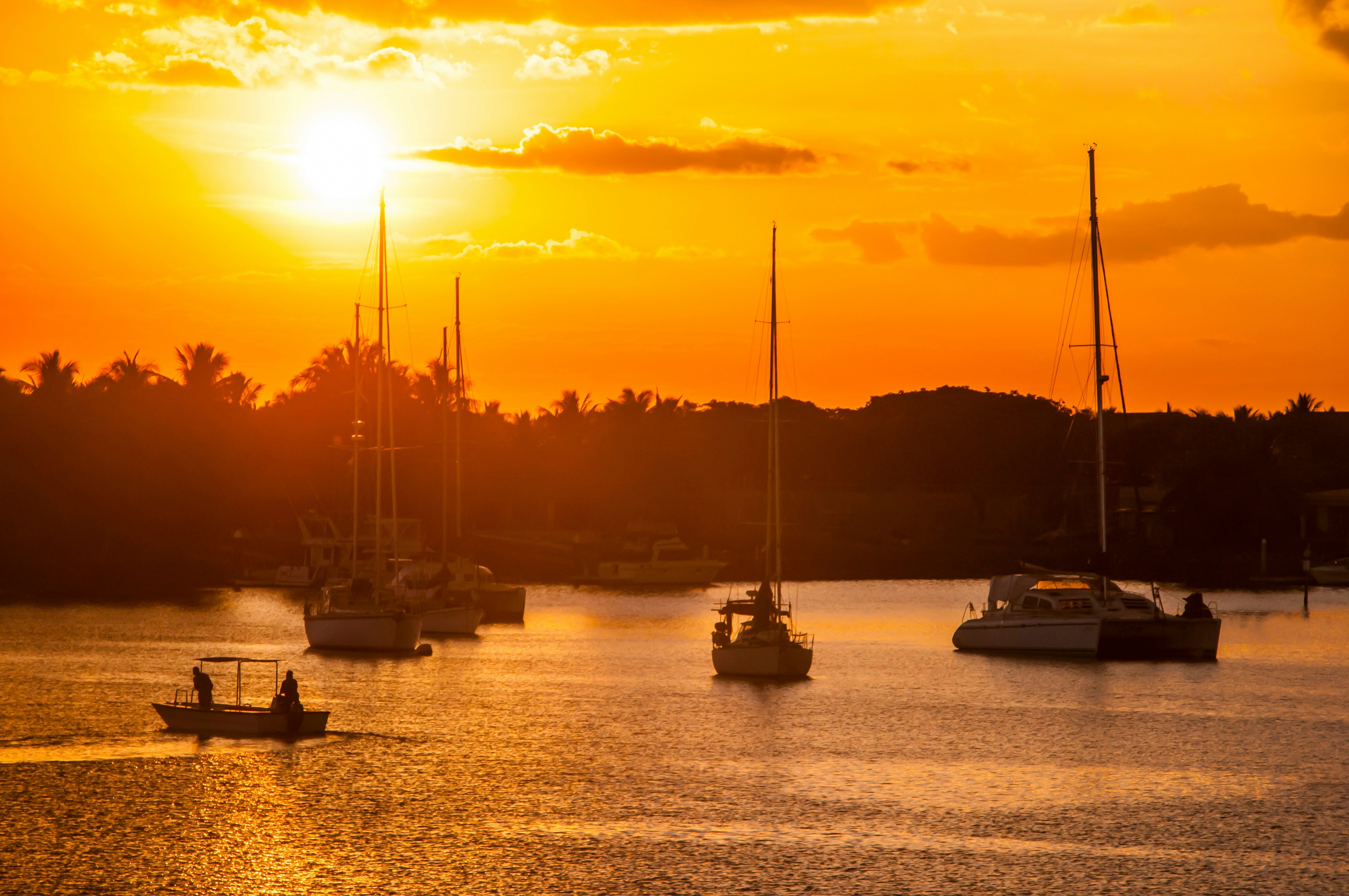 Two people in a small fishing boat and larger yachts are in silhouette as the sun sets casting an orange glow over a harbor.