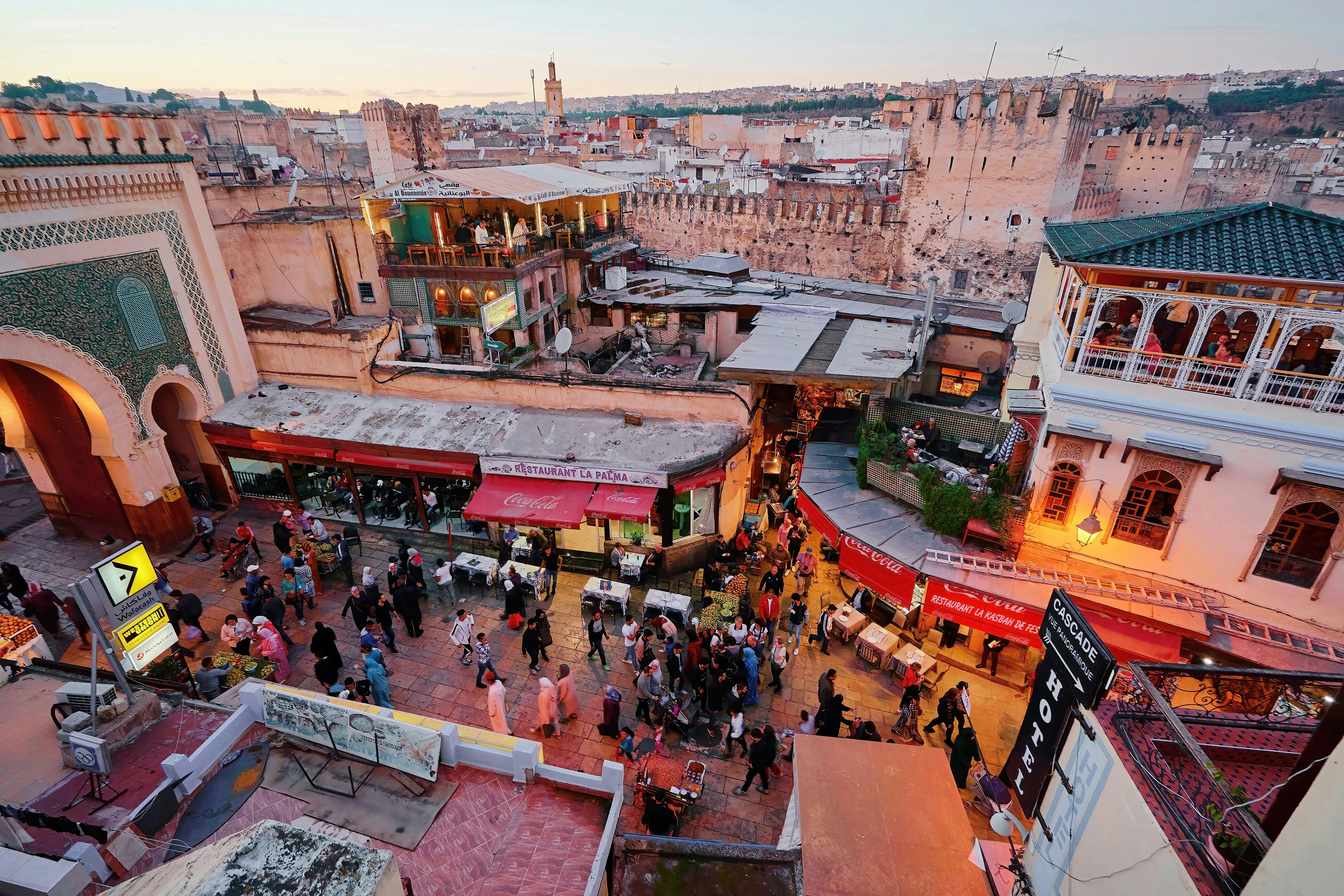 People wander down streets and push carts through an ancient medina. Rooftop bars and ancient walls dominate the skyline.