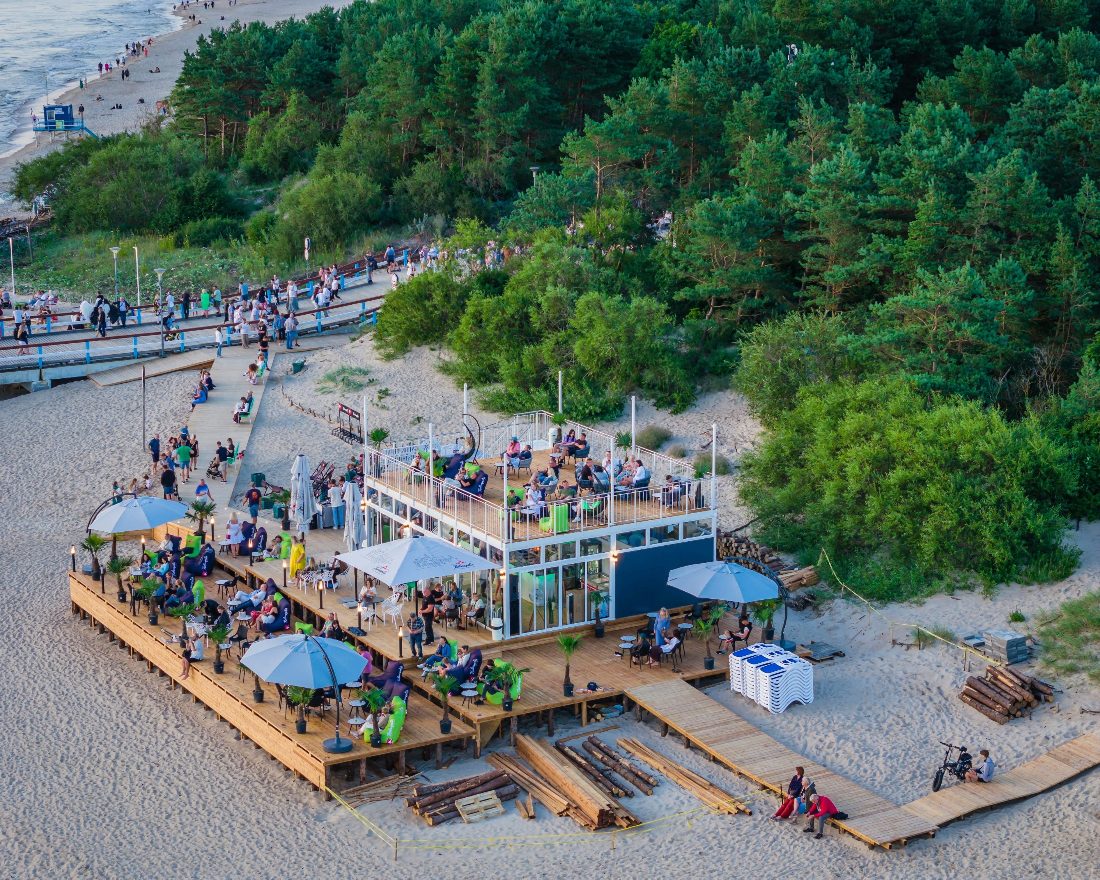 A beach bar with decking backed by woodland. Patrons sit on comfortable chairs.