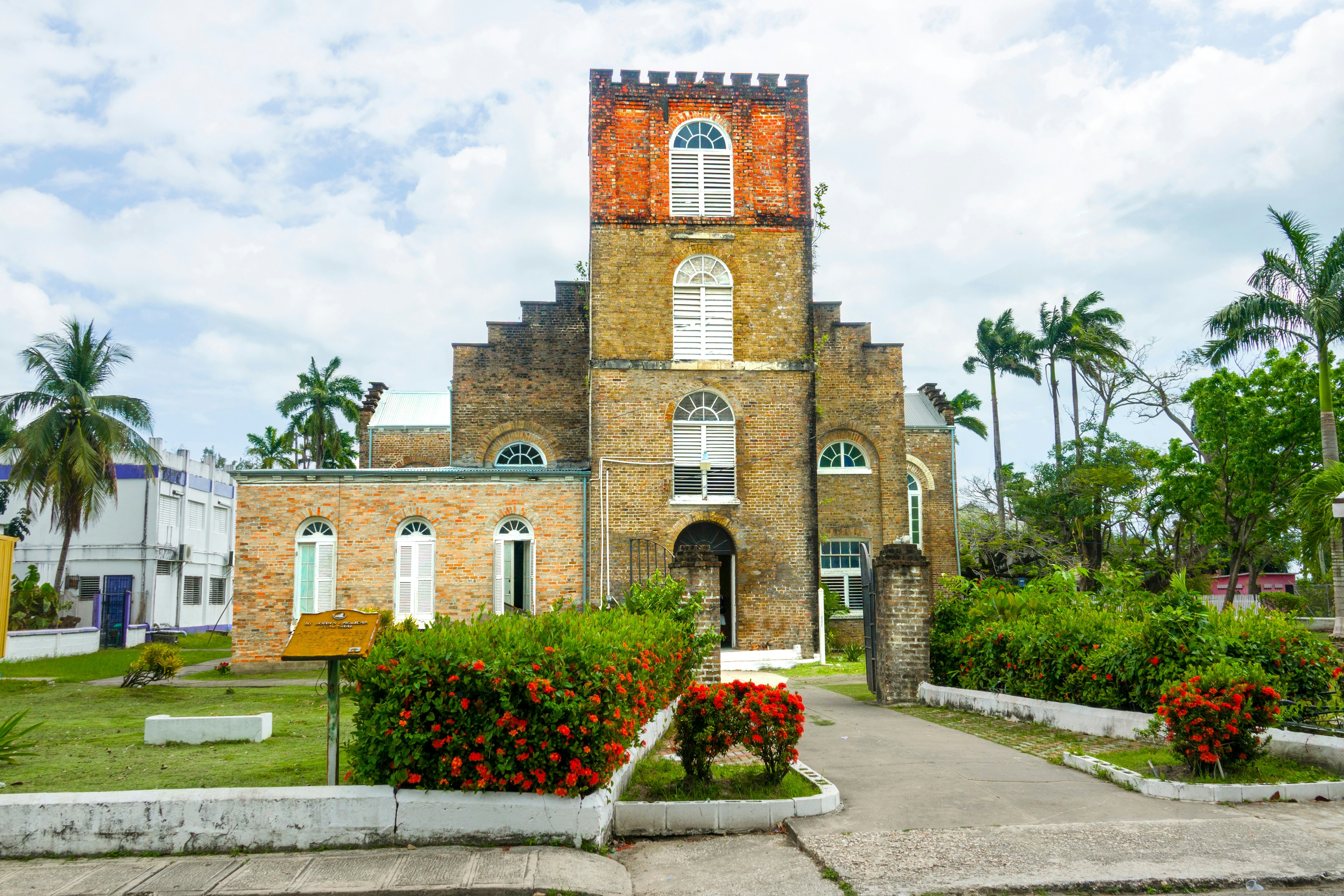 Exterior of a brick churck with palm trees in the background and red-flowered bushes in front on a bright, cloudy day.