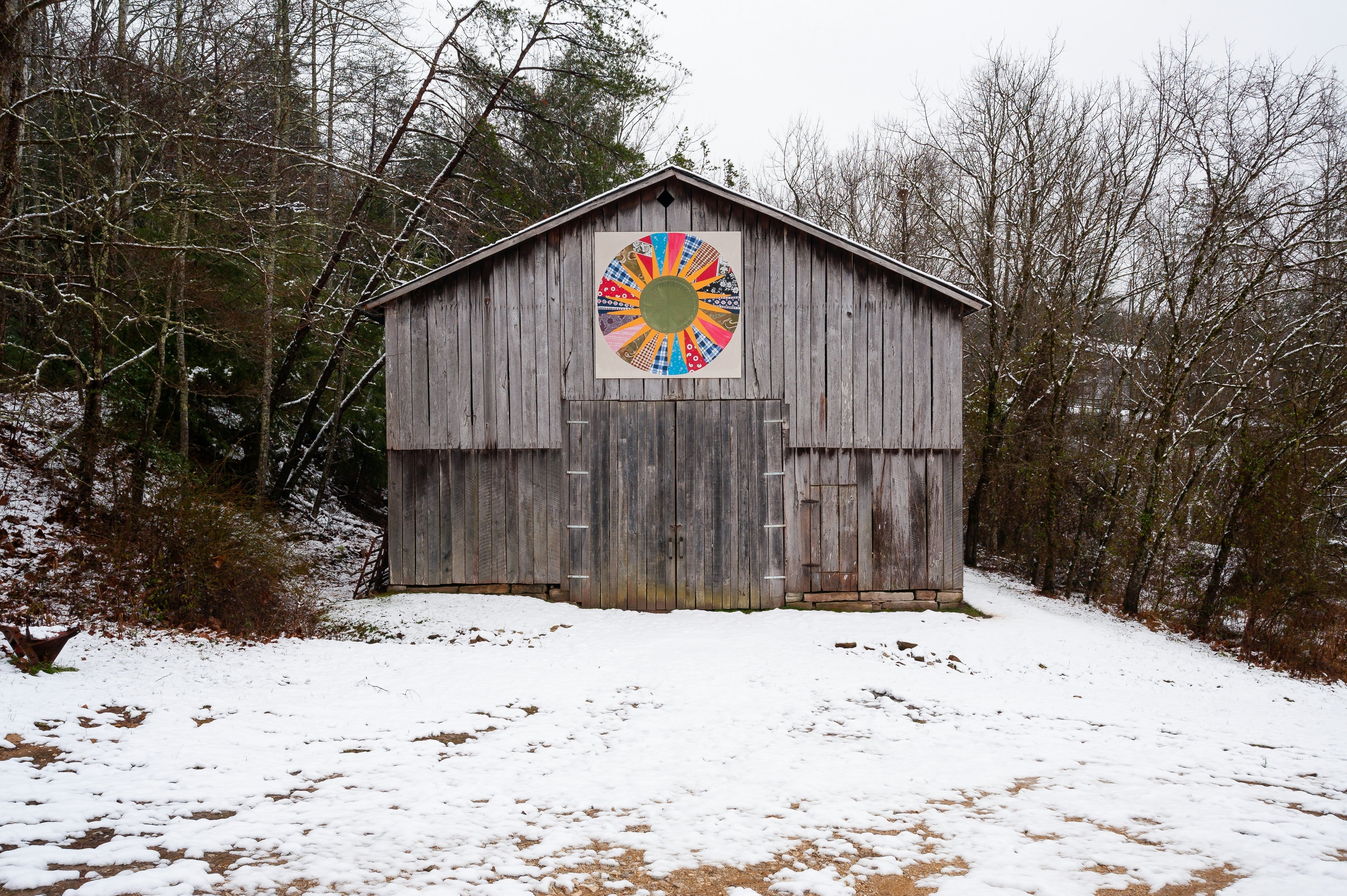 A snowy scene of a rustic farm barn with a quilt print atop the main door on an overcast day.